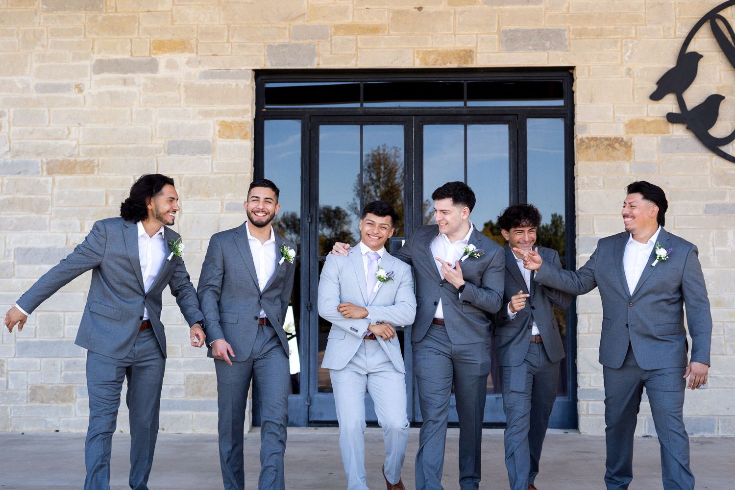 Groomsmen in matching gray suits laughing and posing together outside a stone building.