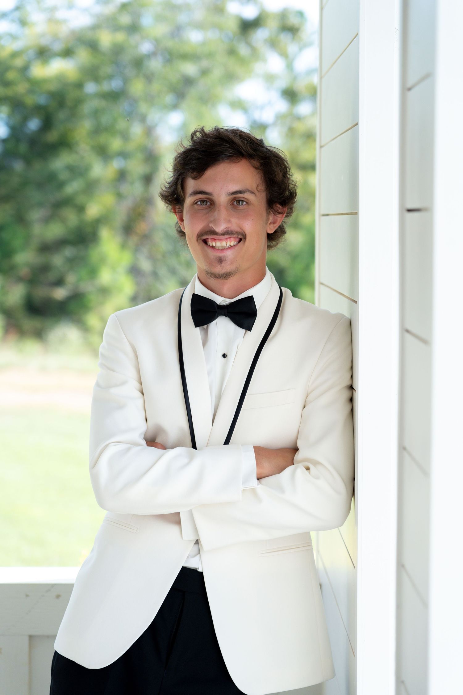A person in a classic white tuxedo jacket with black bow tie smiles while posing against white columns outdoors.