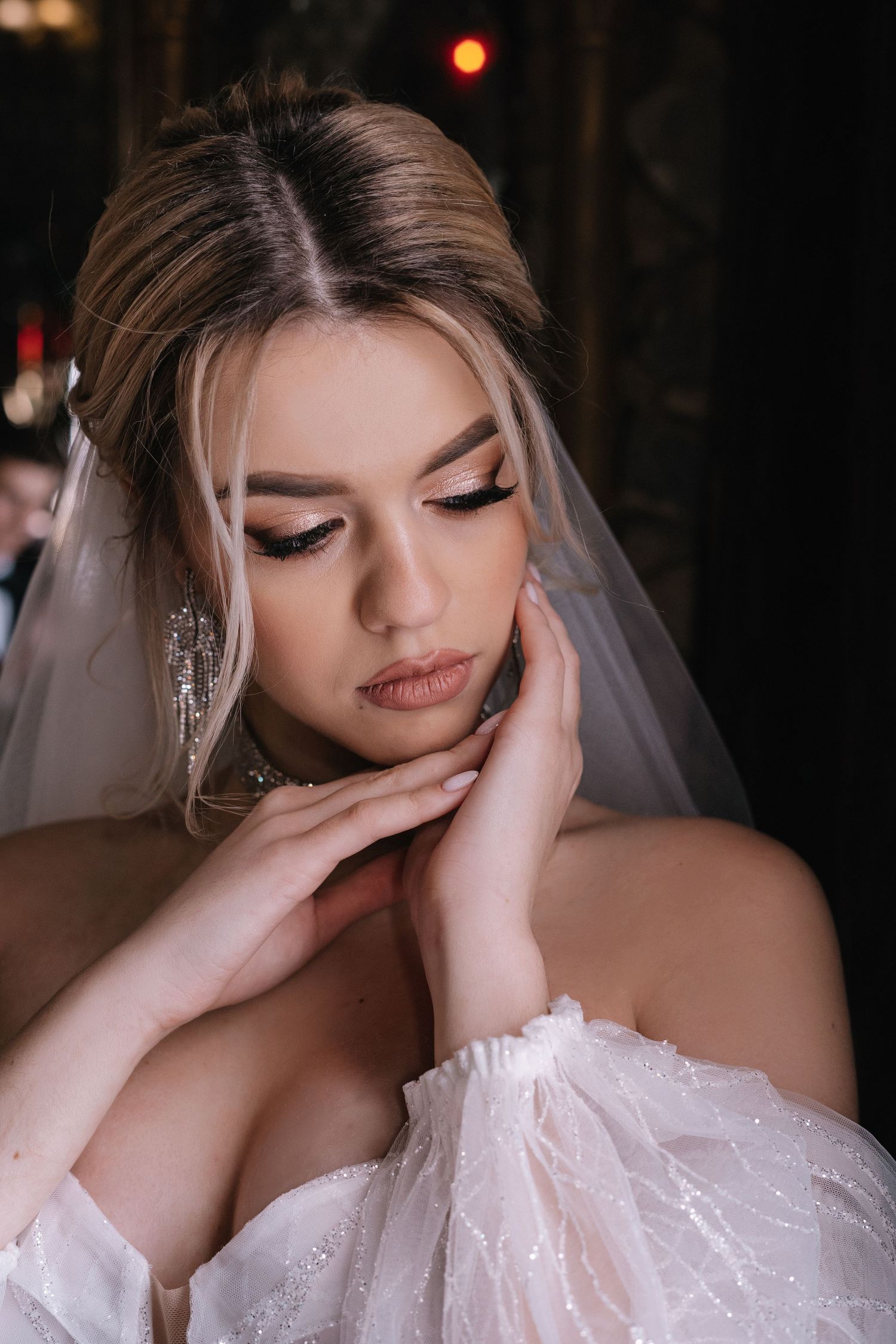 A bride in a white wedding dress with sparkling details wears elegant makeup and dangling earrings in a moody portrait.