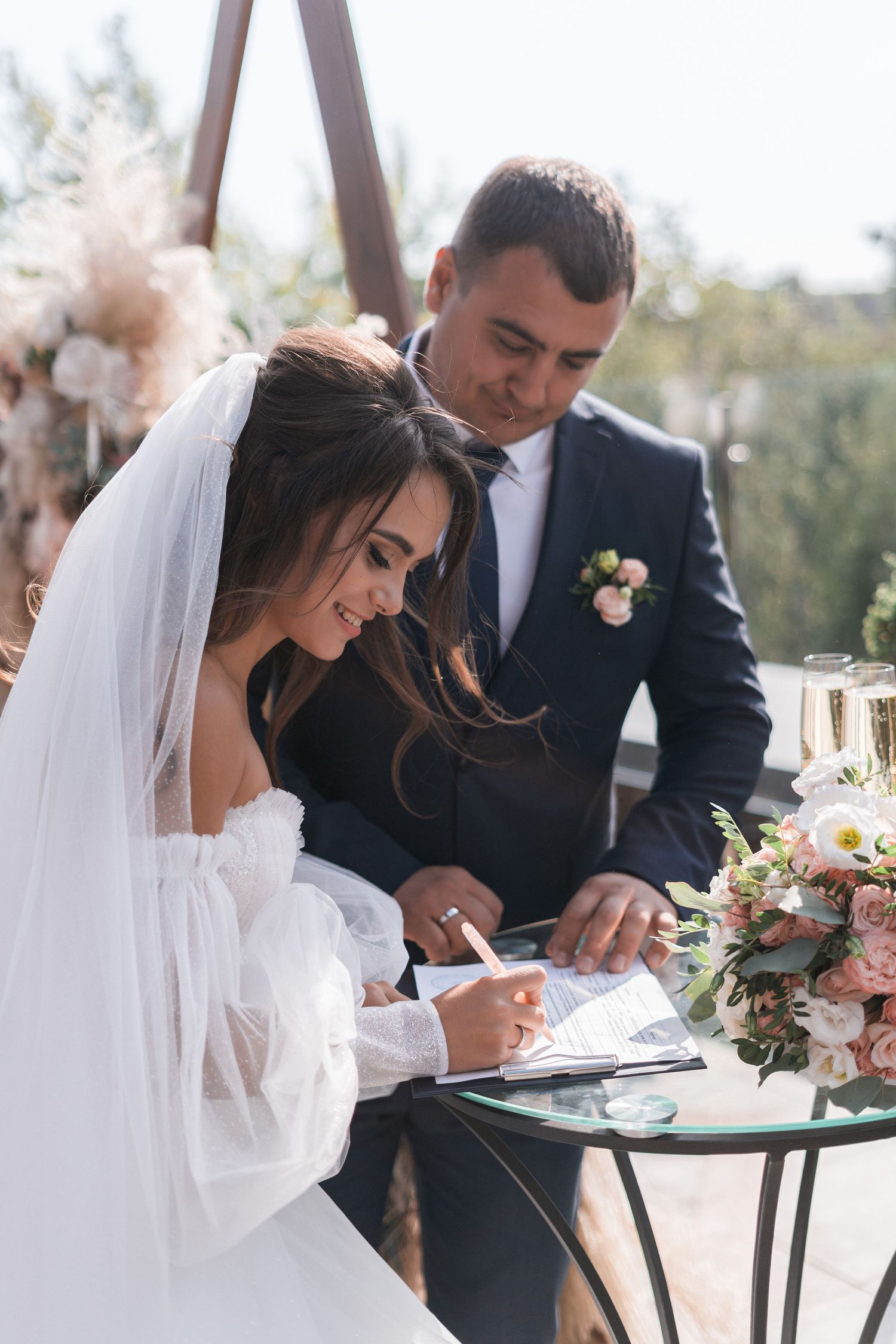 Couple signing marriage documents at an outdoor wedding ceremony with floral decorations.