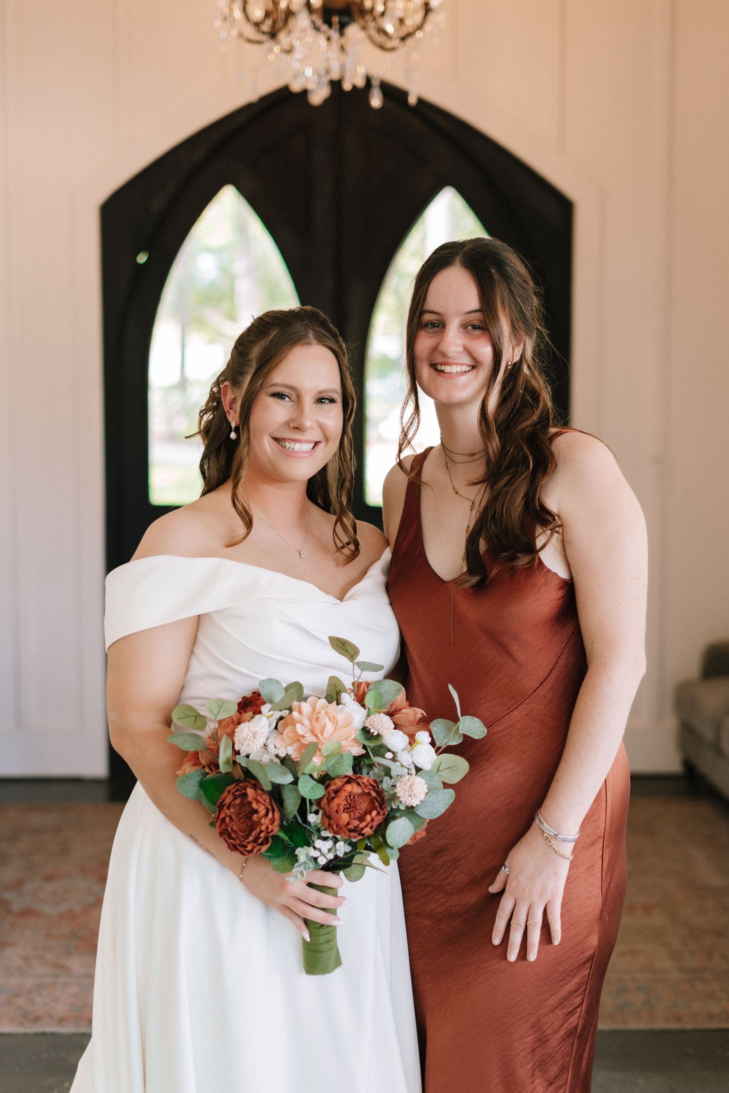 Wedding party members in rust-colored and white dresses pose together with floral bouquets in an elegant indoor venue.