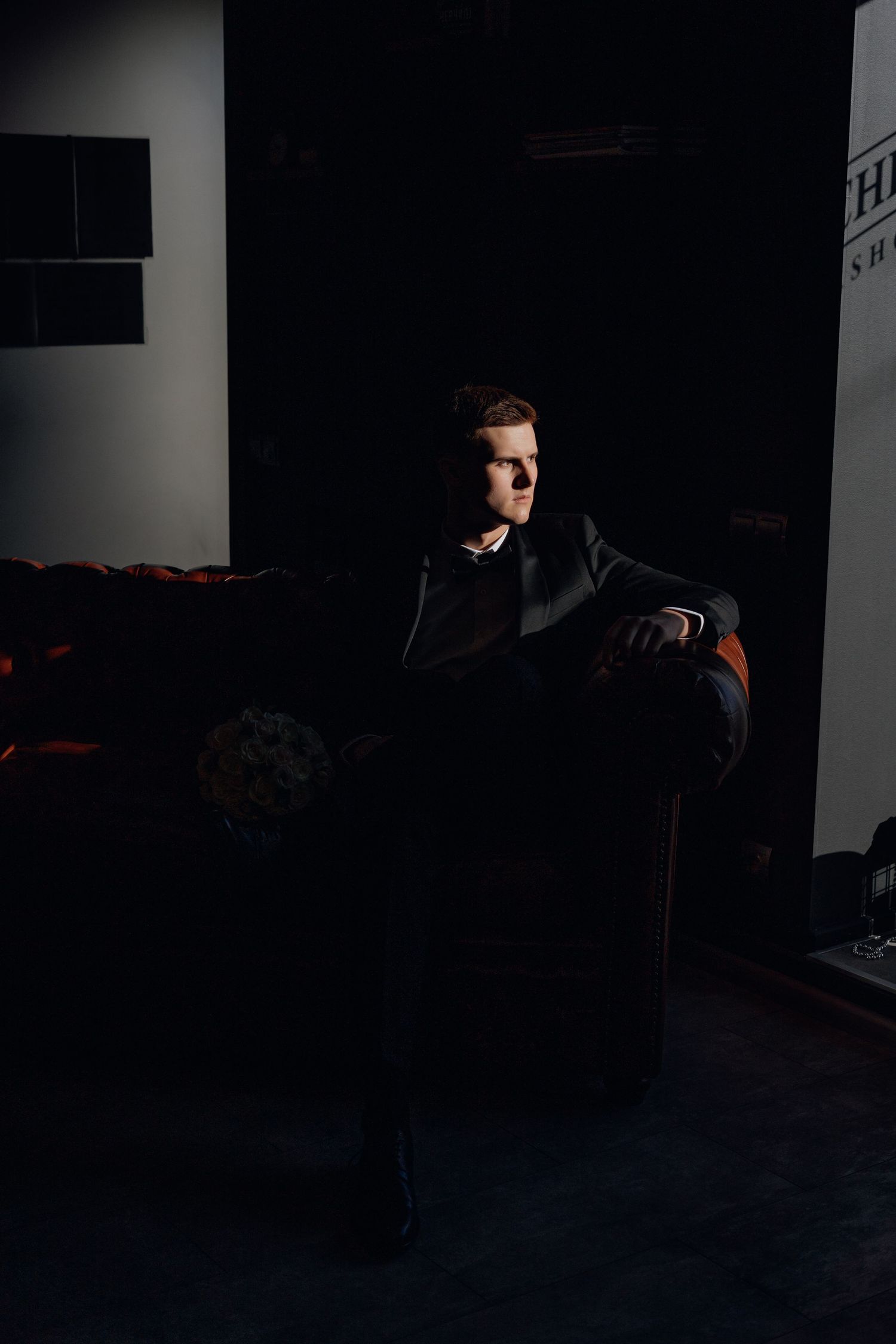 A dark and moody portrait in low-key lighting shows a figure seated in a vintage leather armchair against a shadowy background.
