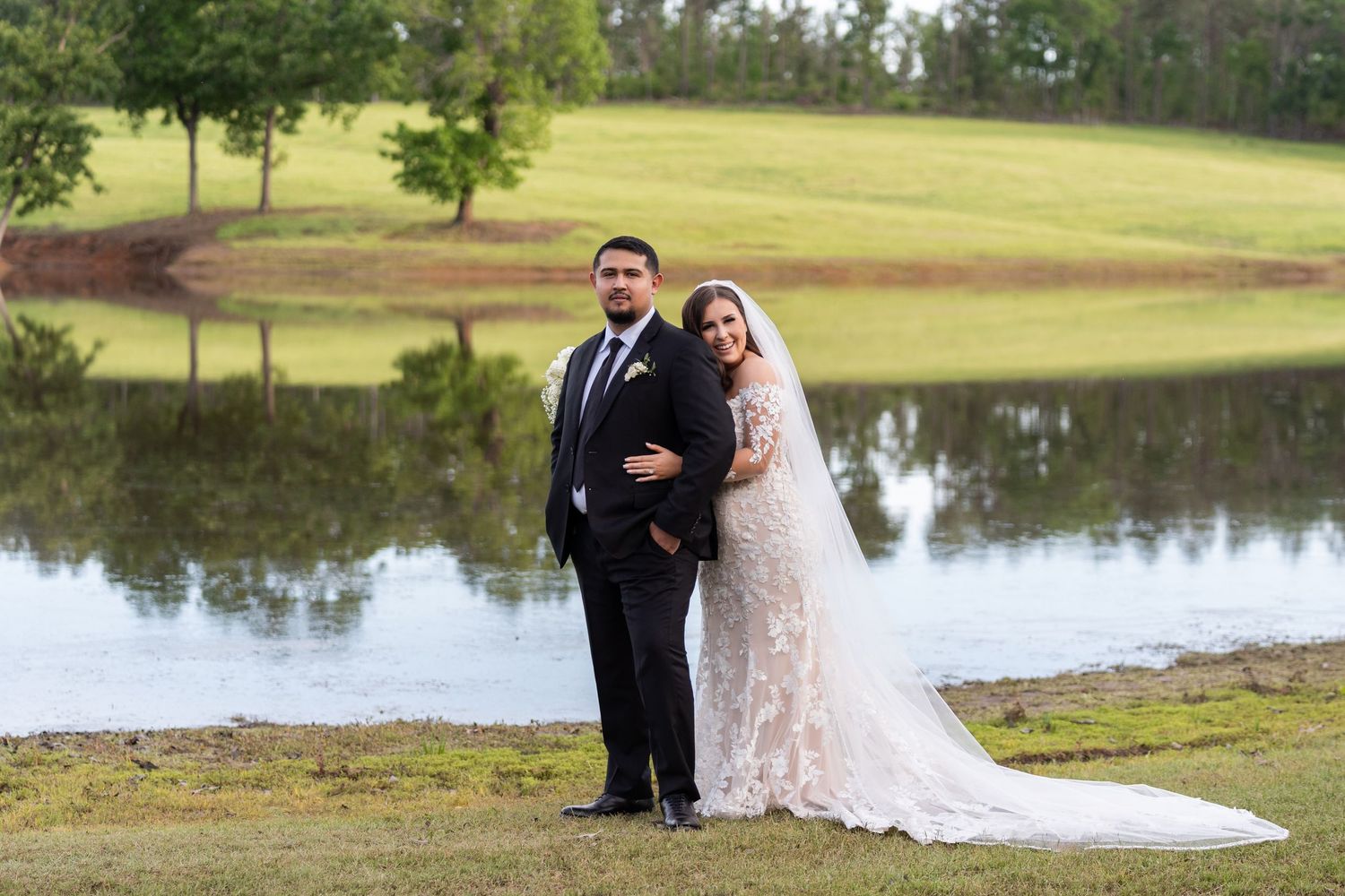 A couple in formal wedding attire poses together near a scenic lake surrounded by trees and greenery.