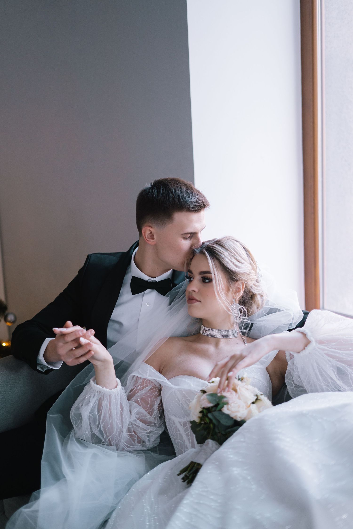Elegant bride and groom share intimate moment on couch in formal wedding attire with white bouquet.
