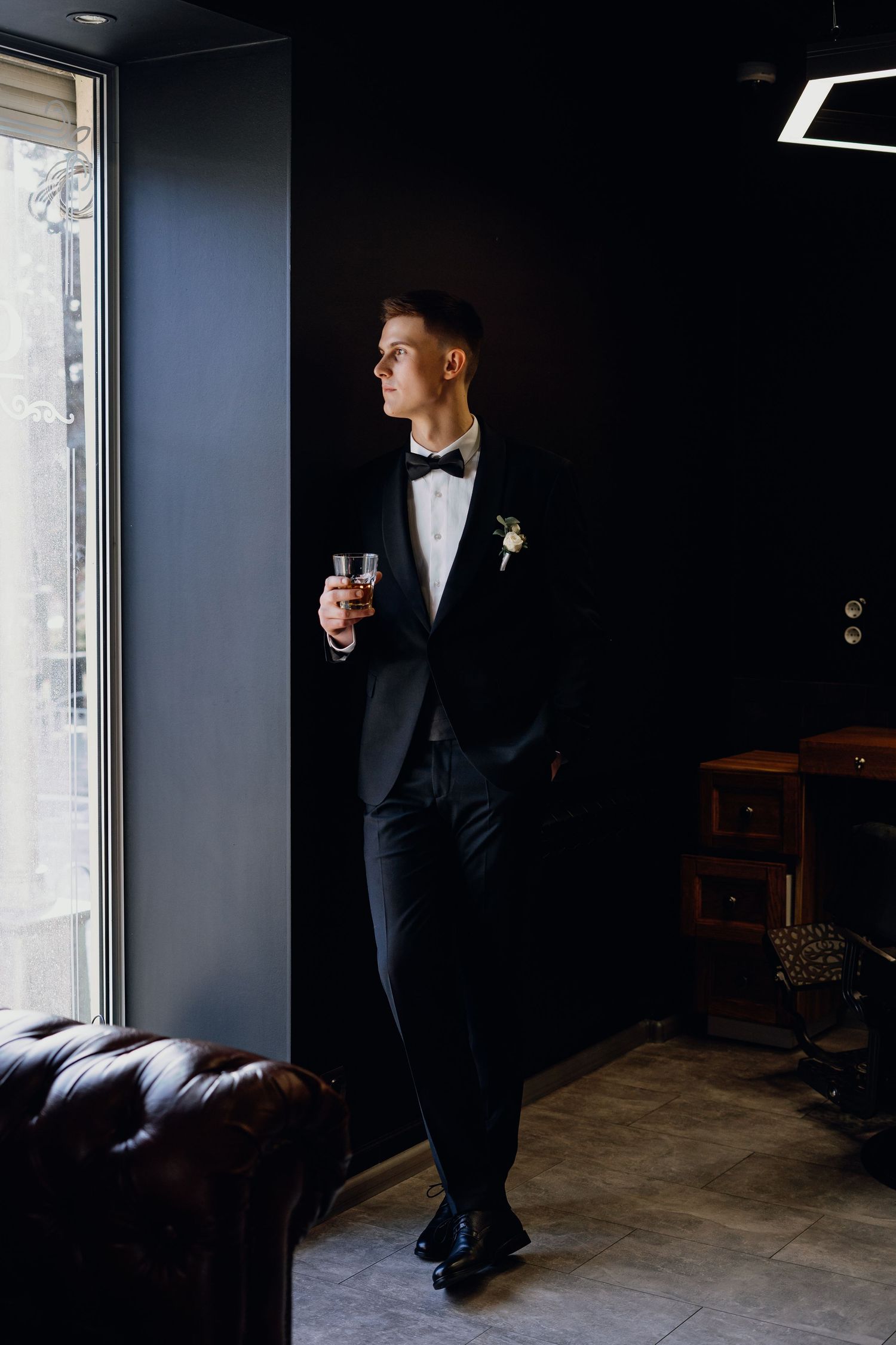 A groom in a black tuxedo stands in dramatic lighting near a window creating a moody and elegant wedding portrait.
