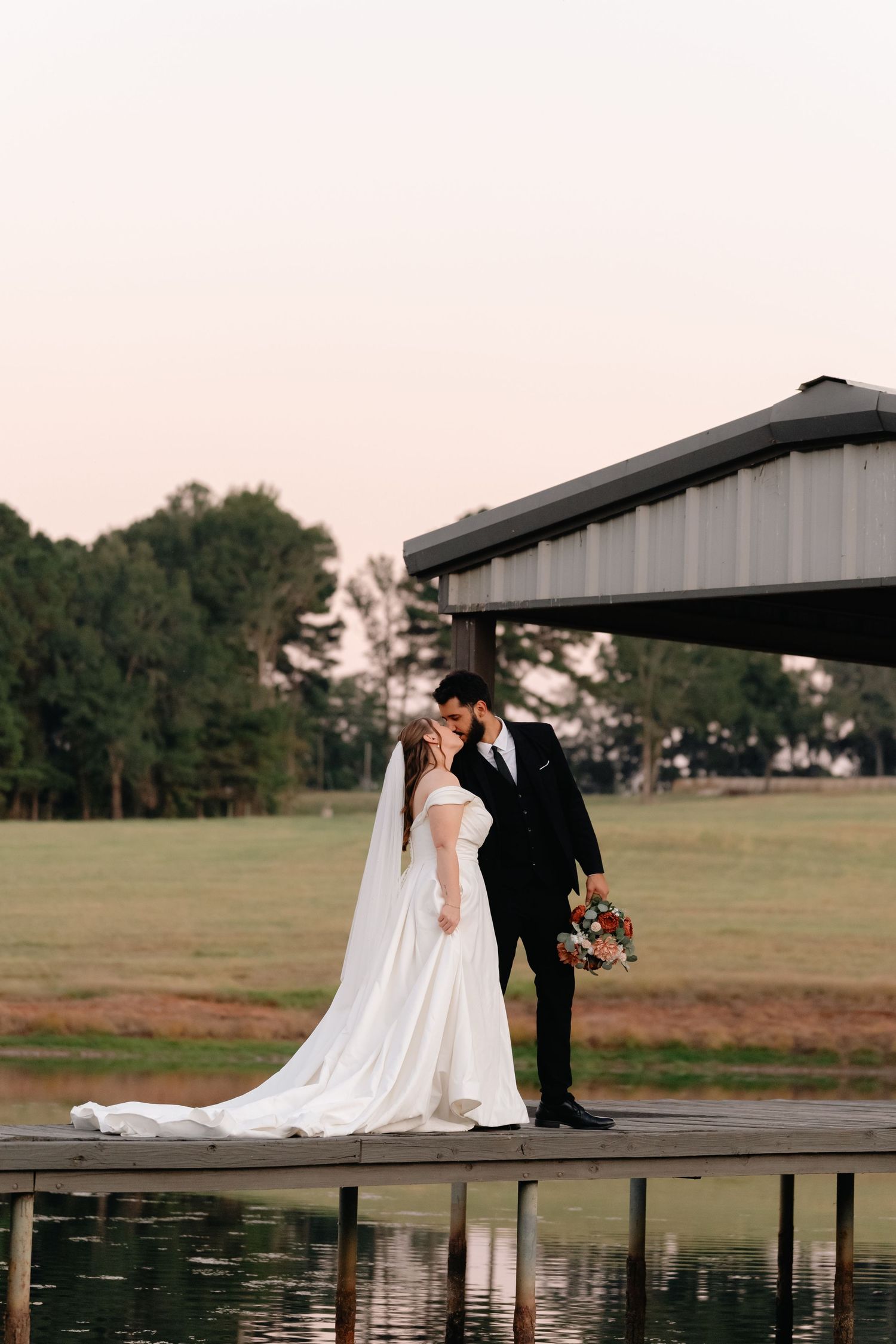 A wedding couple poses on a wooden dock by a lake at dusk with a rustic building in the background.