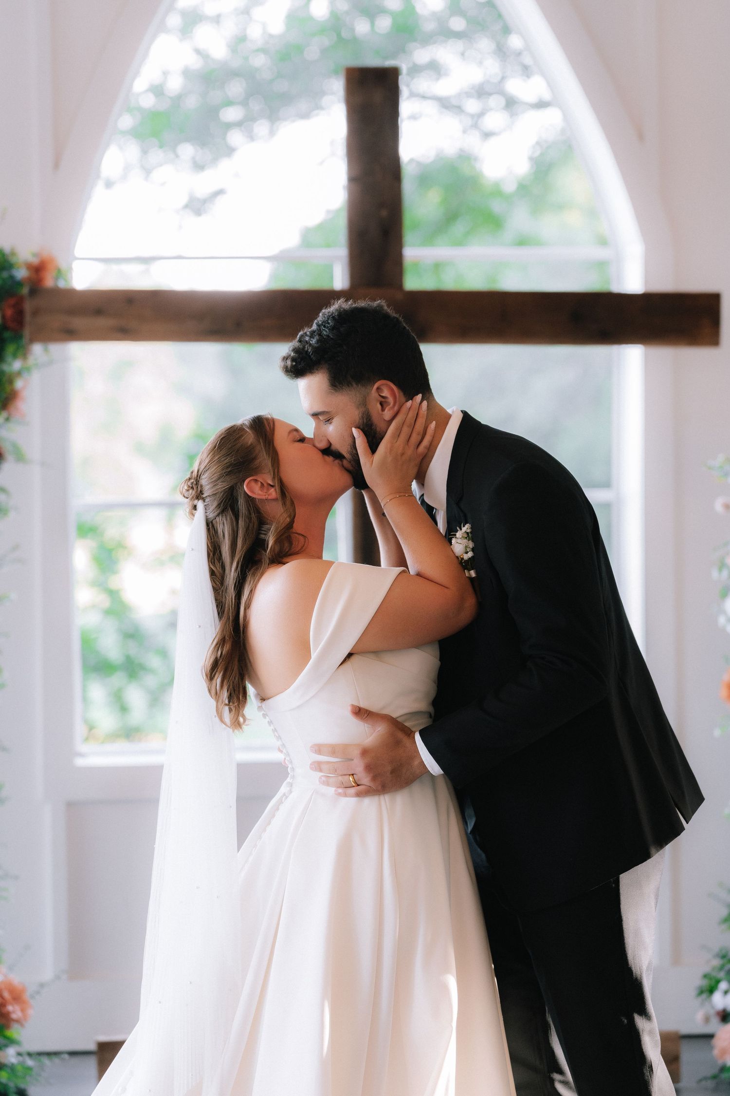 Romantic first kiss moment between newlyweds under arched white colonnade.
