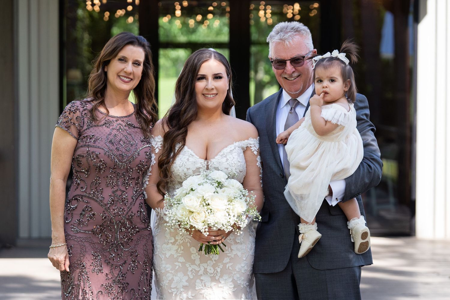 A group stands together at a formal event with a bride in a white lace gown holding a white floral bouquet.