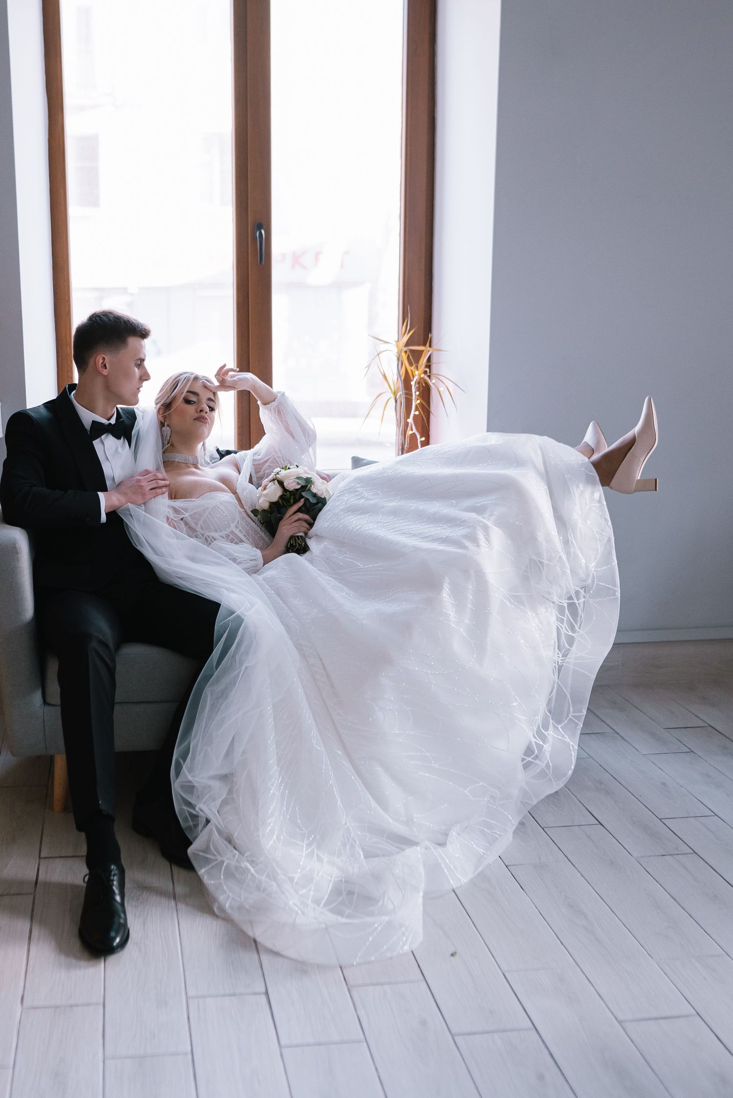 Elegant wedding couple relaxes on grey couch, bride in flowing white dress with feet up in intimate indoor setting.