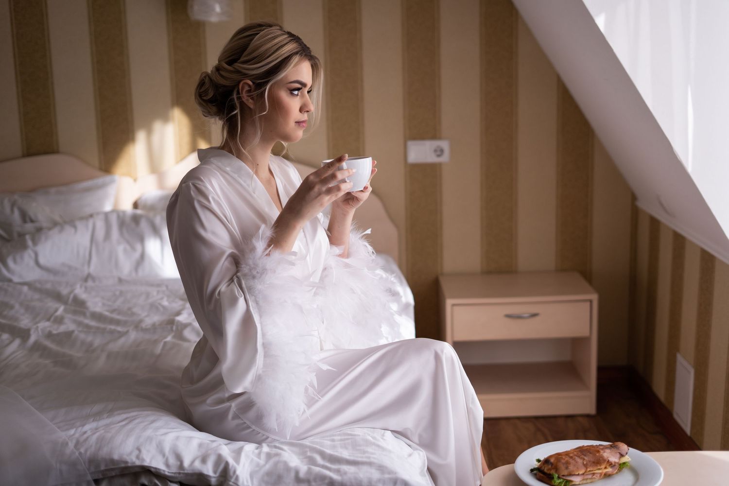 Woman in white robe sits on bed drinking morning coffee with breakfast tray in cozy bedroom interior.