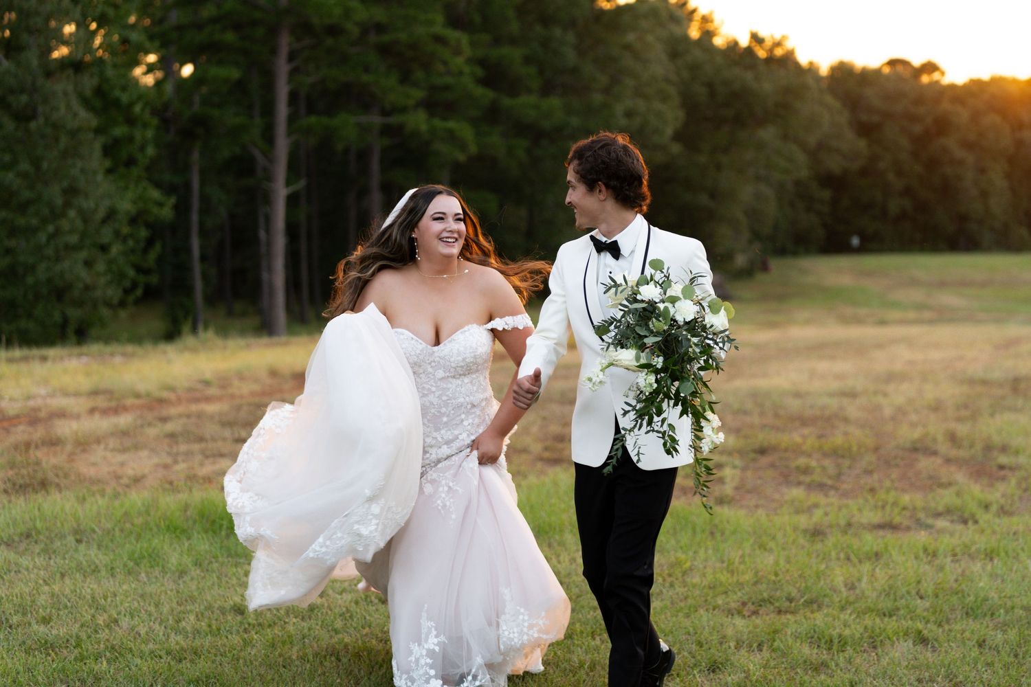 A couple in wedding attire walks together in a grassy field at sunset with trees in the background.