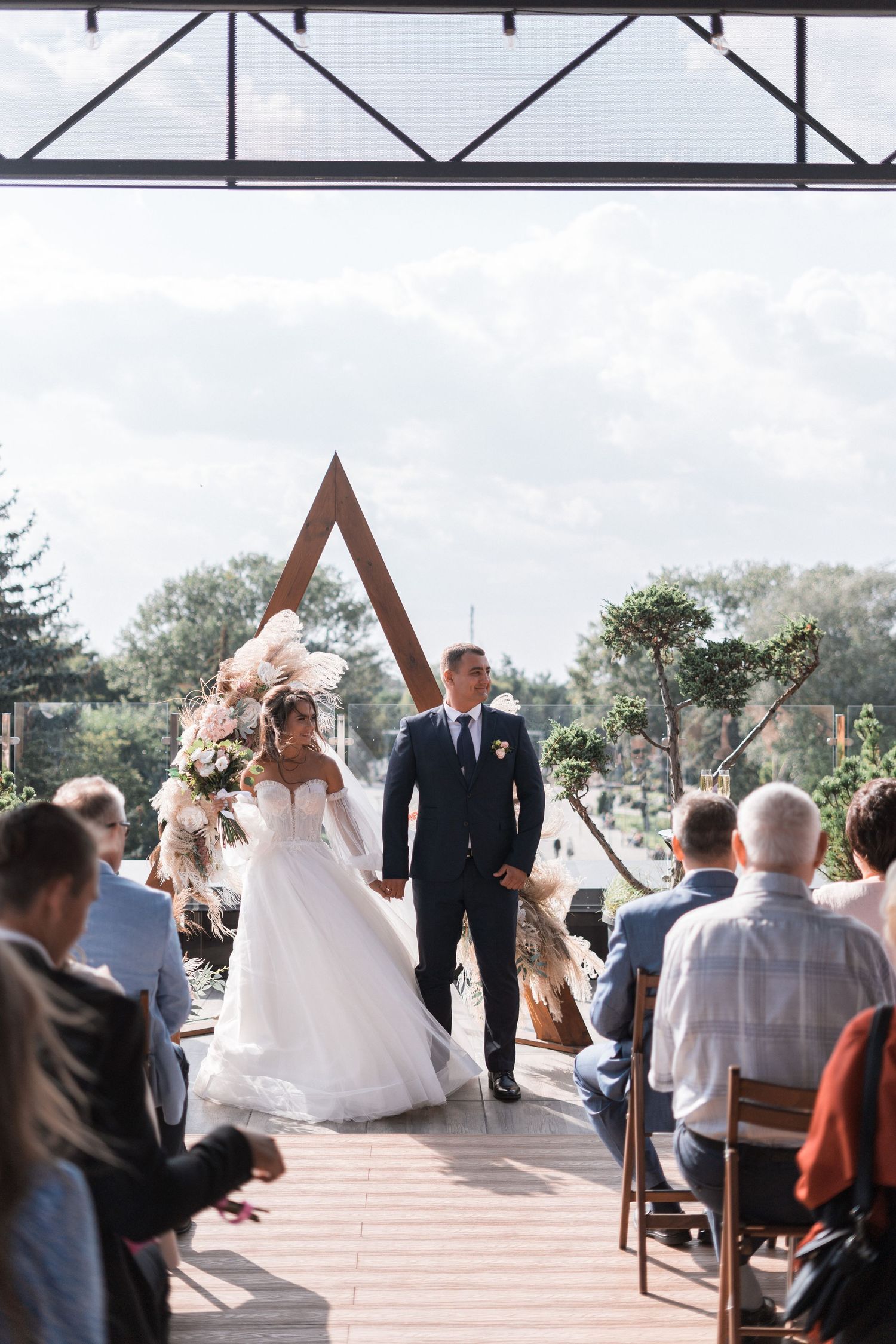 Wedding ceremony taking place outdoors under a wooden triangular arch with guests seated on white chairs.