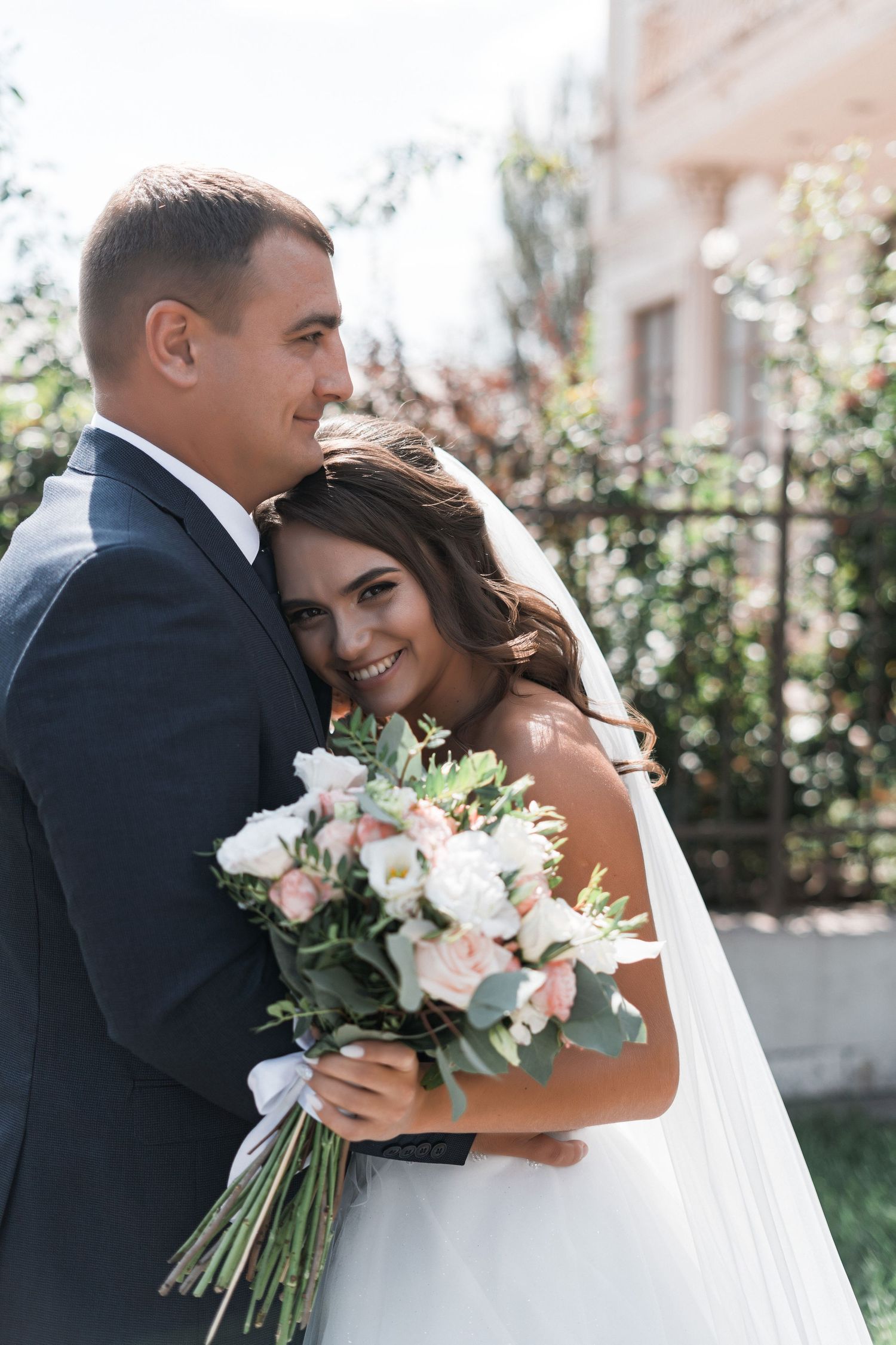 A romantic wedding photo of a couple embracing outdoors, with the bride holding a beautiful bouquet of white and pink flowers.