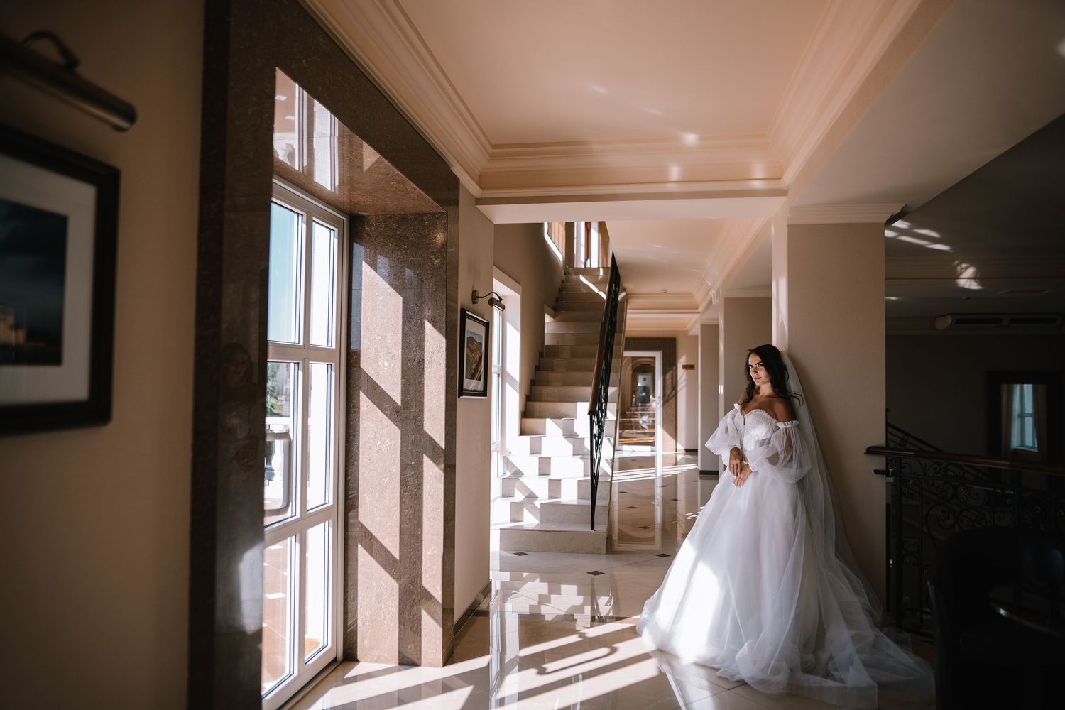 A bride in a white wedding dress stands in a sunlit hallway with large windows casting dramatic shadows on the floor.