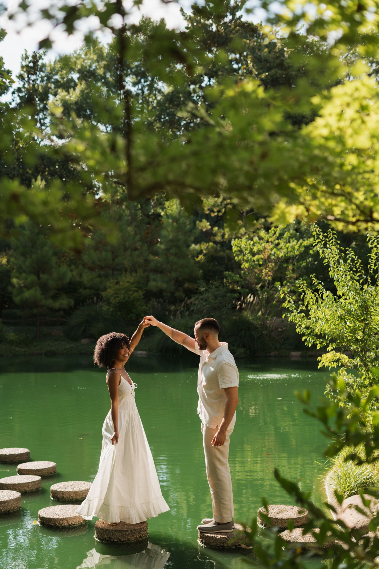 Couple in light summer attire dancing hand in hand on stepping stones across a green pond surrounded by lush trees.