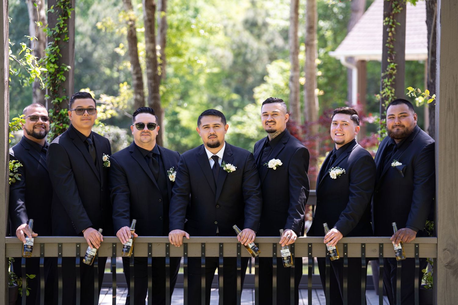 A row of groomsmen in black suits lean against a white railing outdoors with trees in the background.