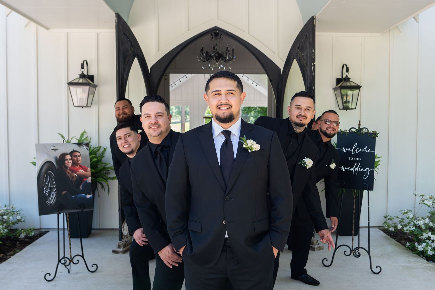 Groomsmen in black suits pose together for formal wedding photos against a white wall with lantern decorations.