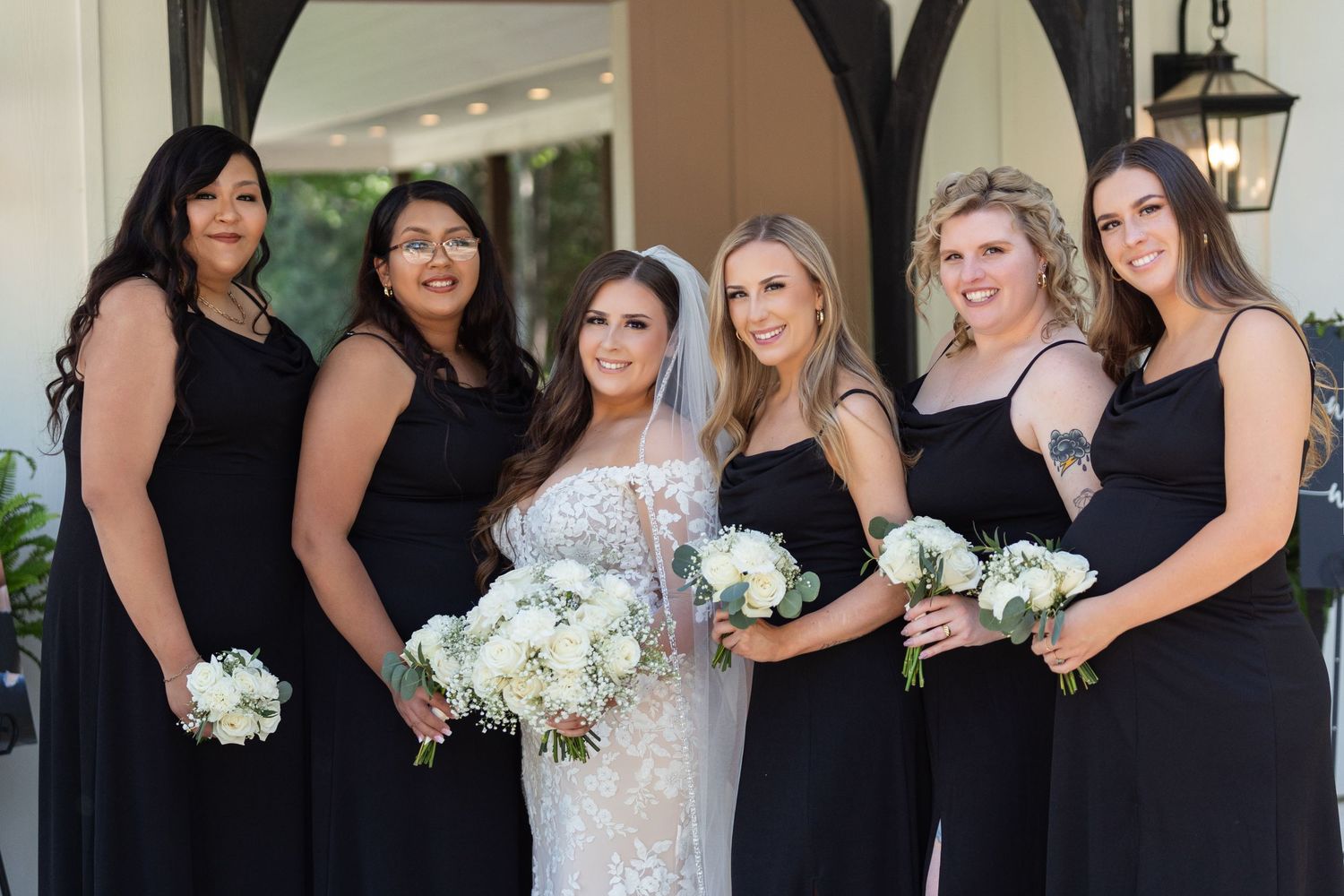 A bridal party in black dresses poses with the bride in white holding white bouquets during a formal wedding.
