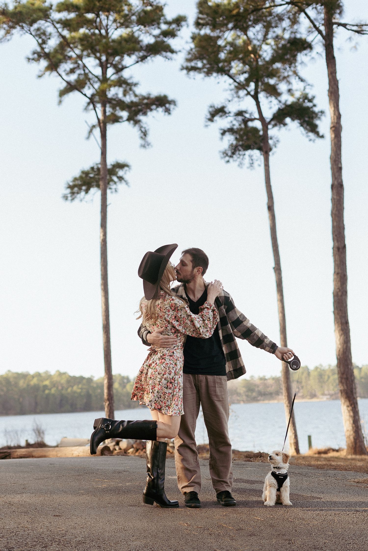 A couple shares a romantic moment with their dog by a lake surrounded by tall pine trees.