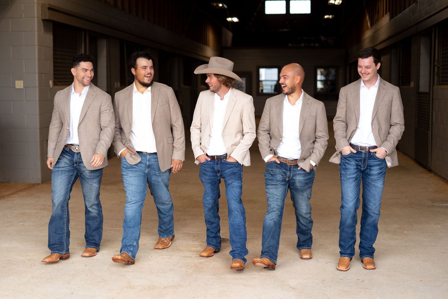 Five groomsmen in matching beige blazers and jeans stand together in a rustic indoor setting.