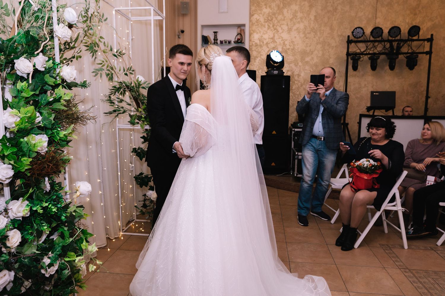 An intimate wedding ceremony moment captured with guests seated on chairs beside a floral-decorated white arch.