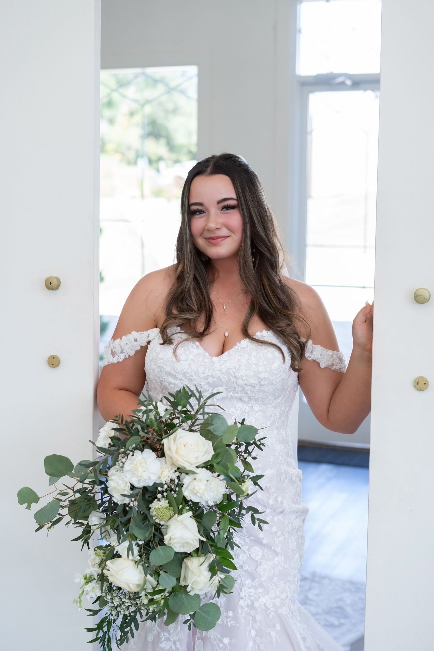 A bride in a lace off-shoulder wedding dress holds a beautiful white and green bouquet with eucalyptus and roses.
