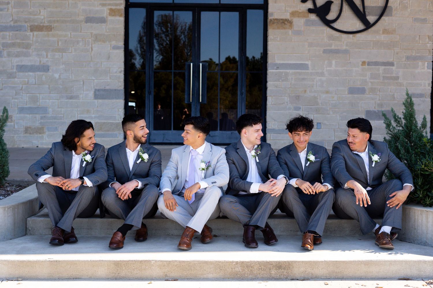Groomsmen in dark suits sit casually on outdoor benches for wedding photos.