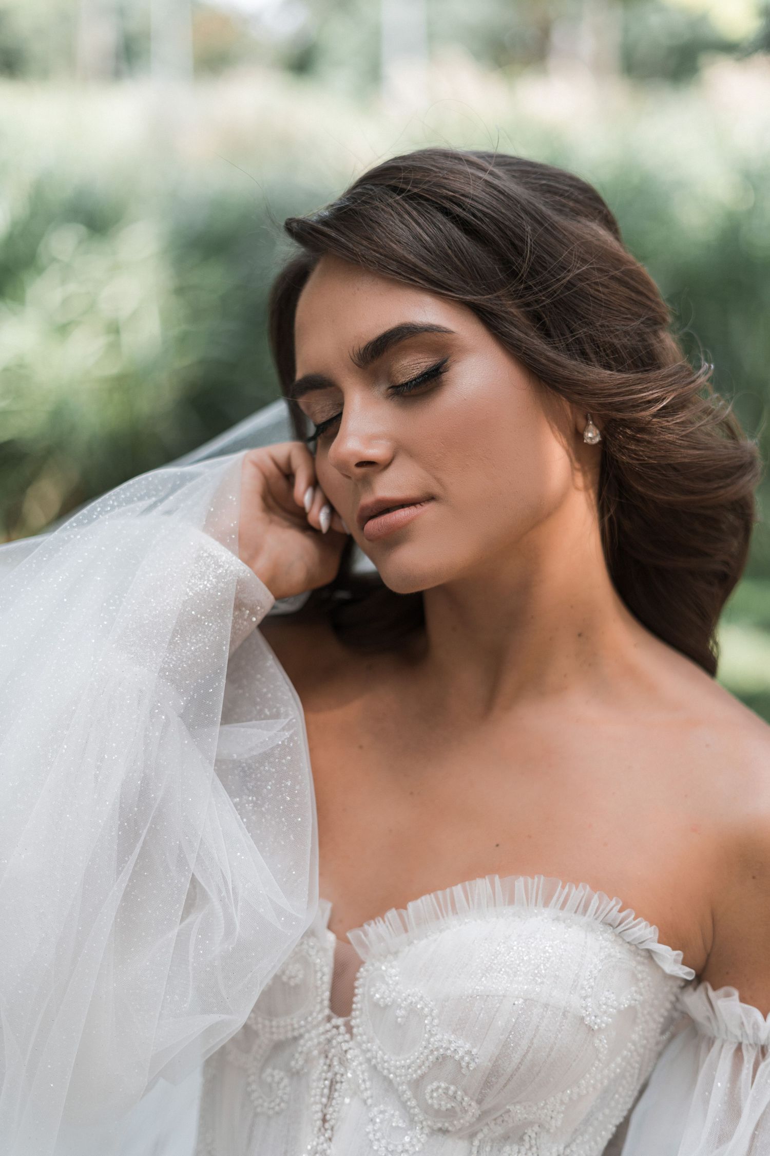 A bride in an off-shoulder lace wedding dress poses with her veil in a romantic outdoor setting.