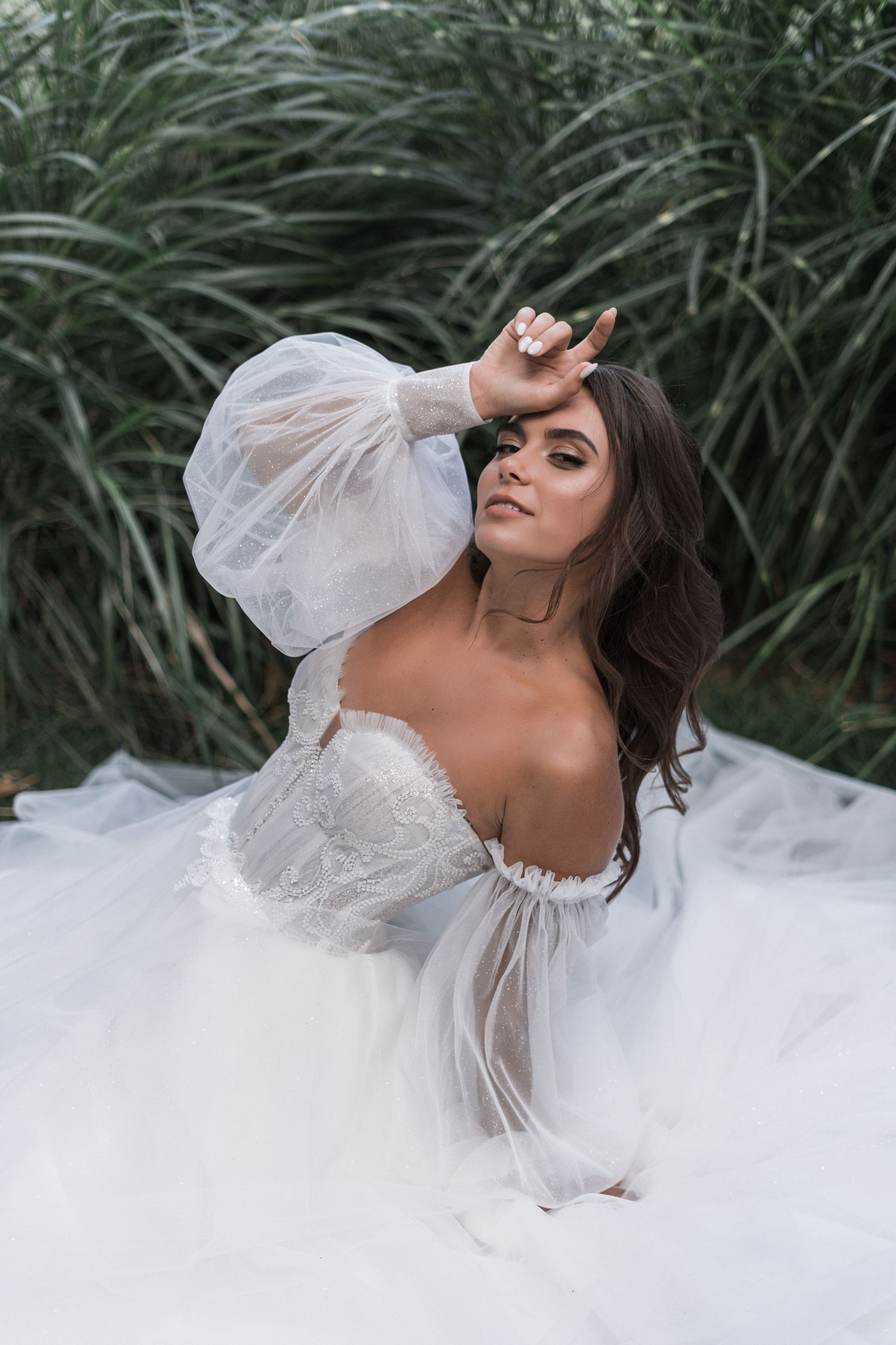 A bride in a flowing white dress poses dramatically against tall grass in an artistic outdoor photoshoot.