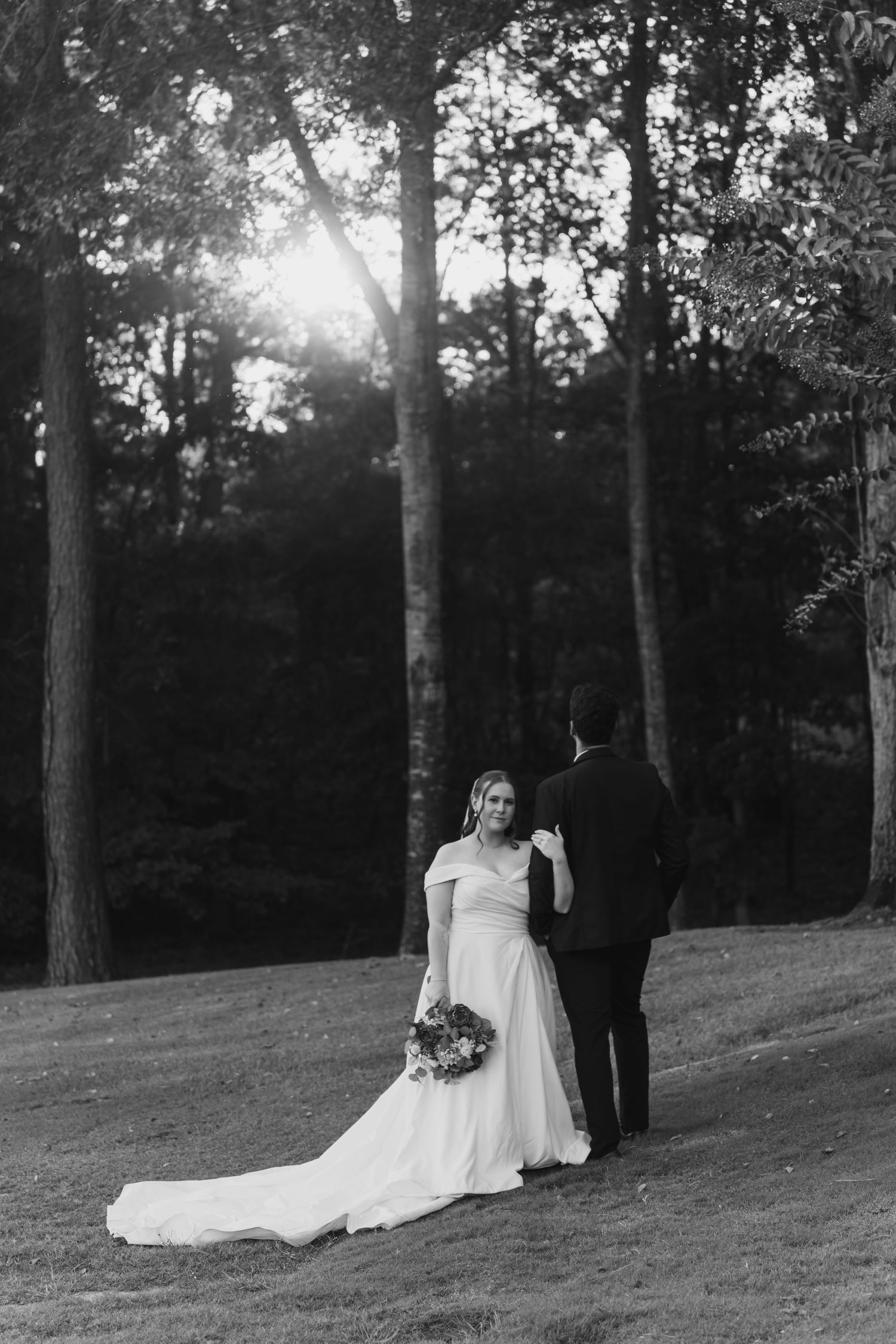 Black and white wedding photos captured on a grassy hillside surrounded by trees at sunset.