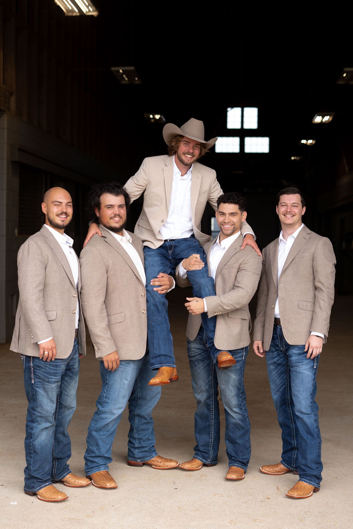Groomsmen in matching tan blazers and blue jeans pose together in a rustic indoor setting.