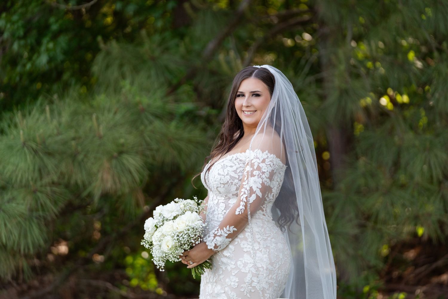 A bride in a lace wedding gown holds a white bouquet while standing in a lush garden setting.