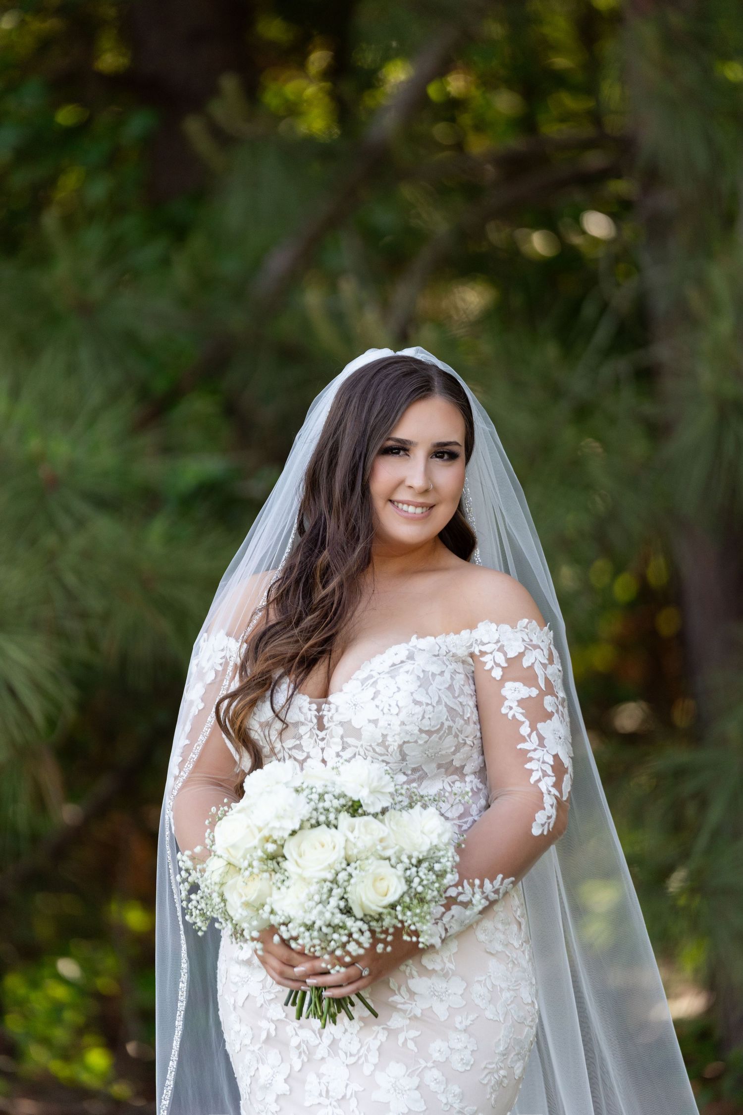 A bride in a white lace wedding dress holds a bouquet of white flowers against a blurred outdoor background.