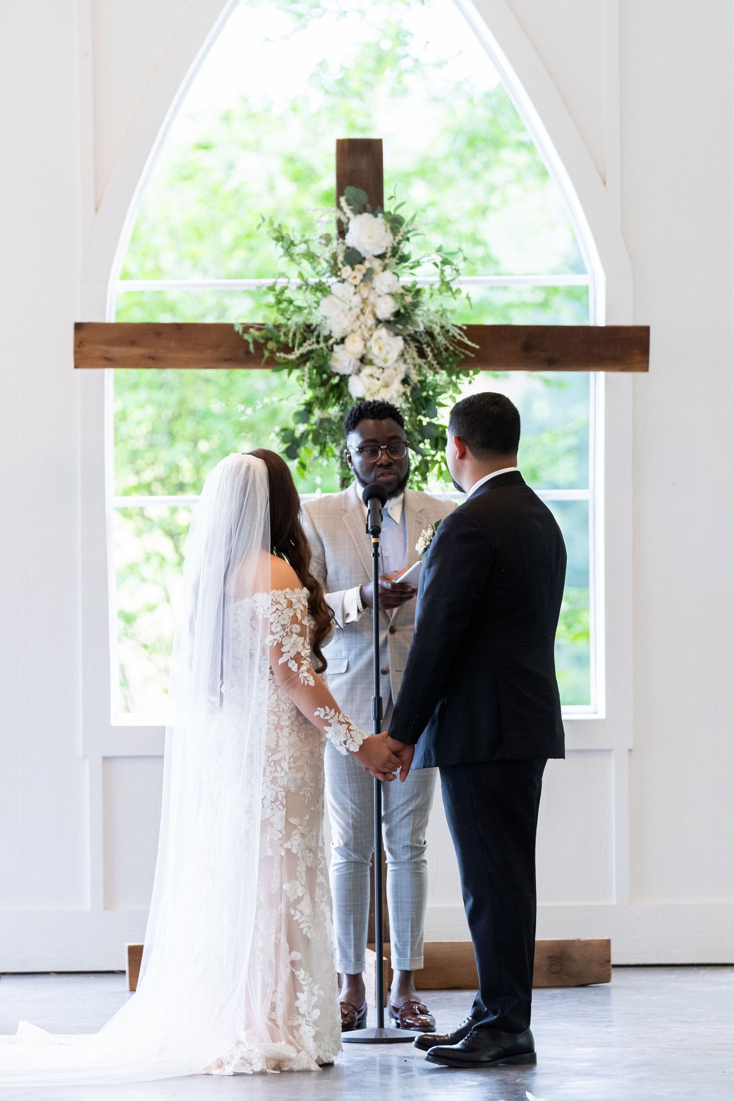 Wedding ceremony in a bright chapel with wooden cross and white floral arrangement under arched window.