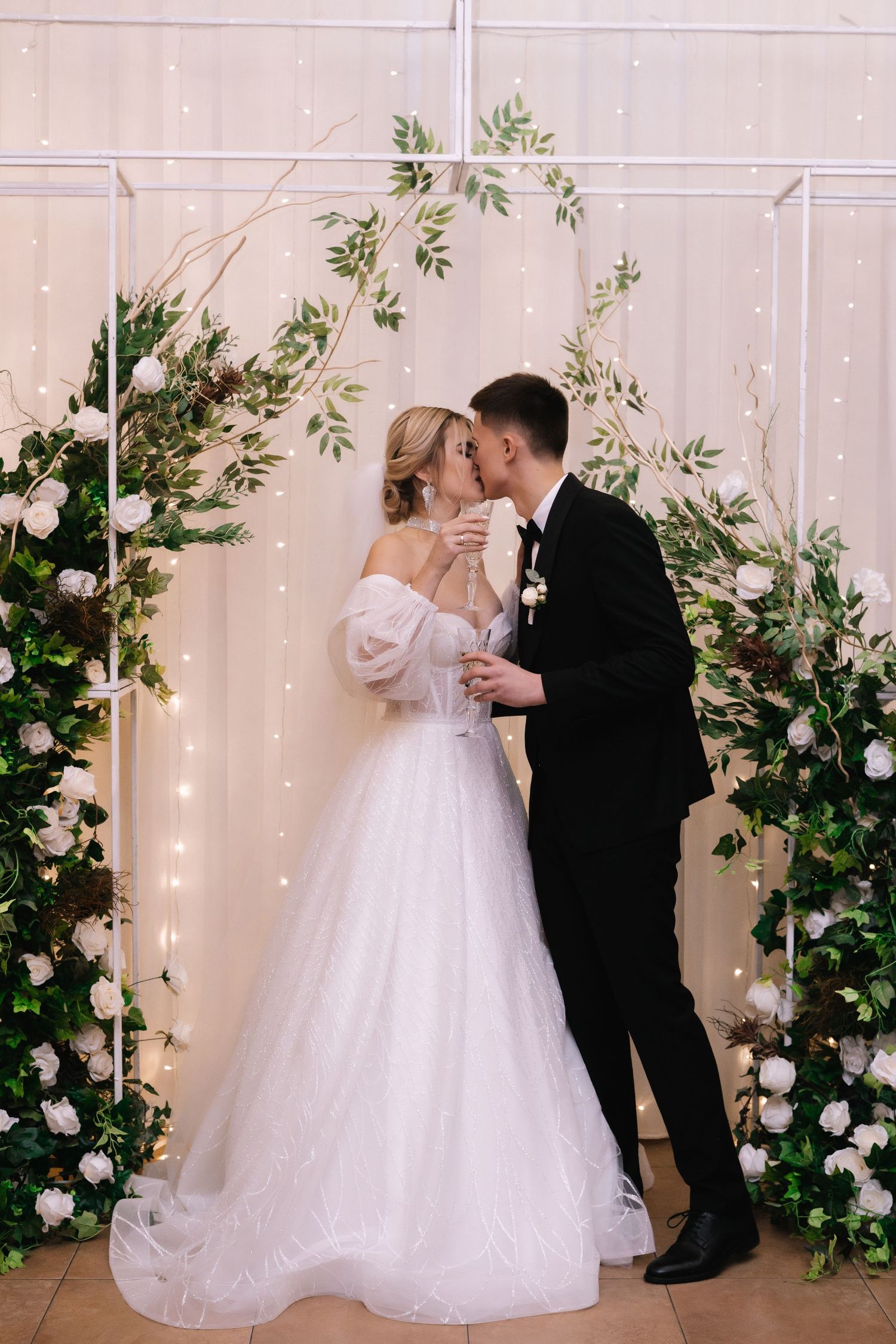 A couple shares their first dance at their wedding surrounded by greenery and twinkling lights on a white backdrop.