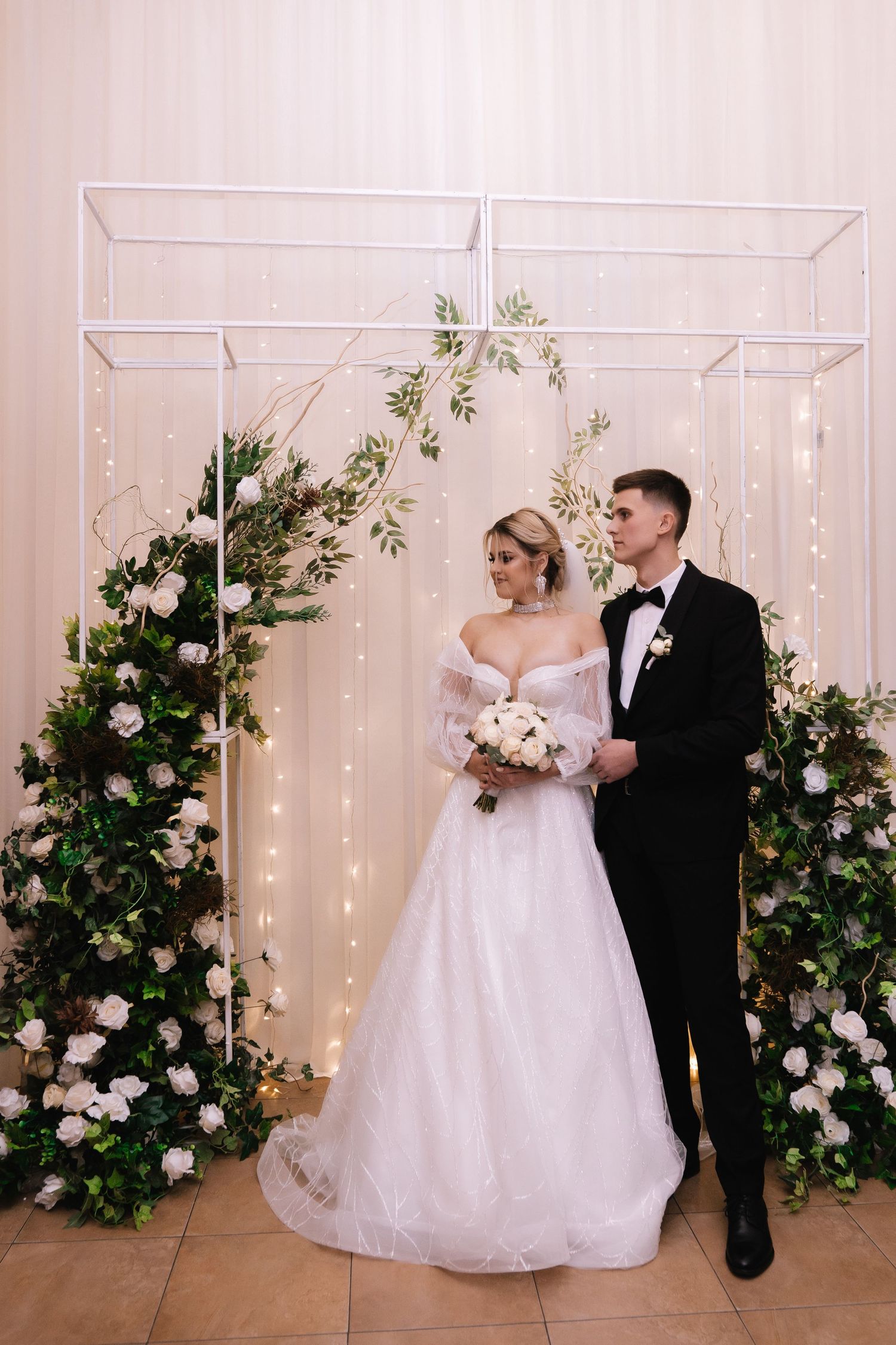 Elegant wedding couple poses under white geometric arch decorated with pink roses and greenery with twinkling lights.
