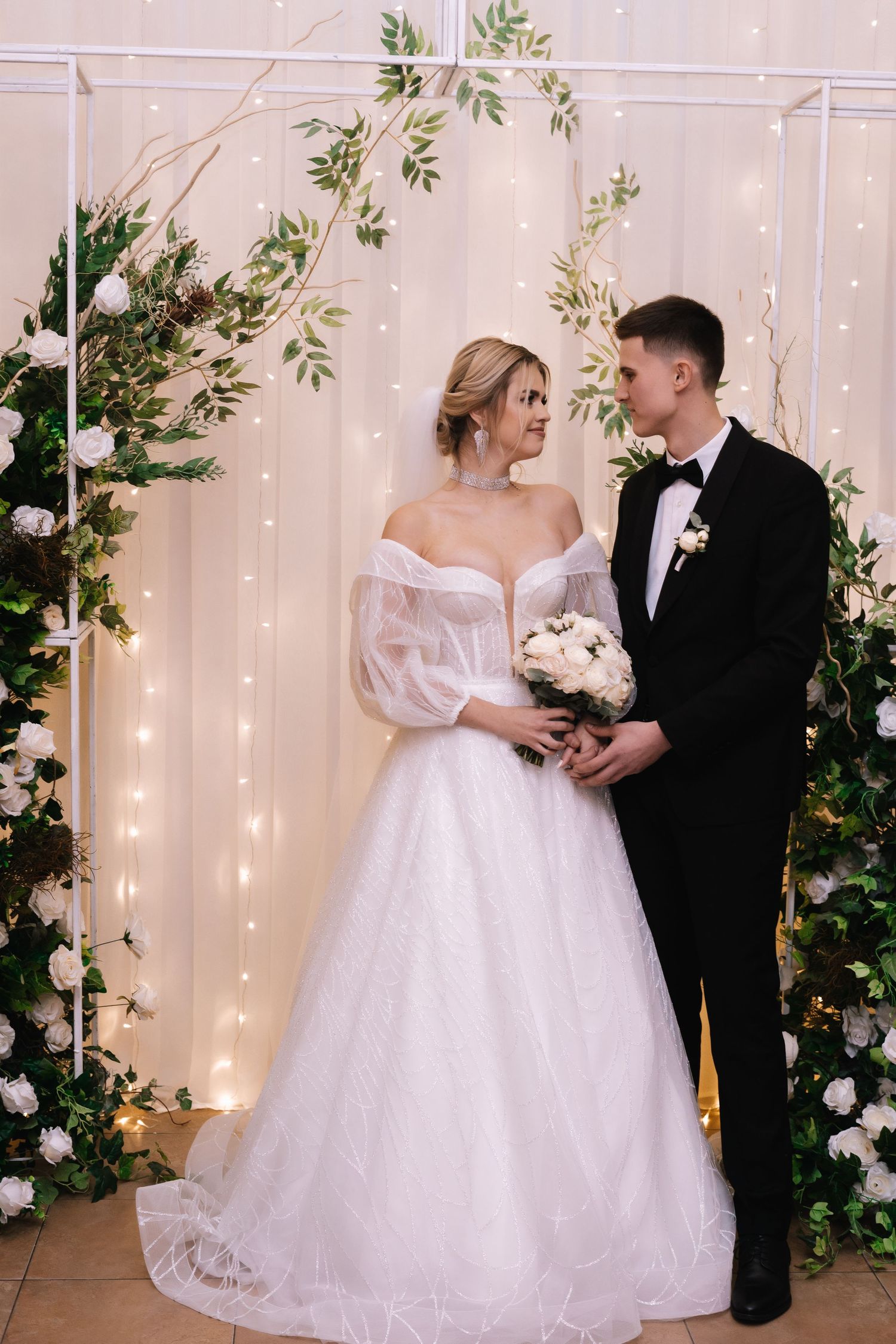 A romantic wedding scene with a couple standing under an arch decorated with greenery and twinkling fairy lights.