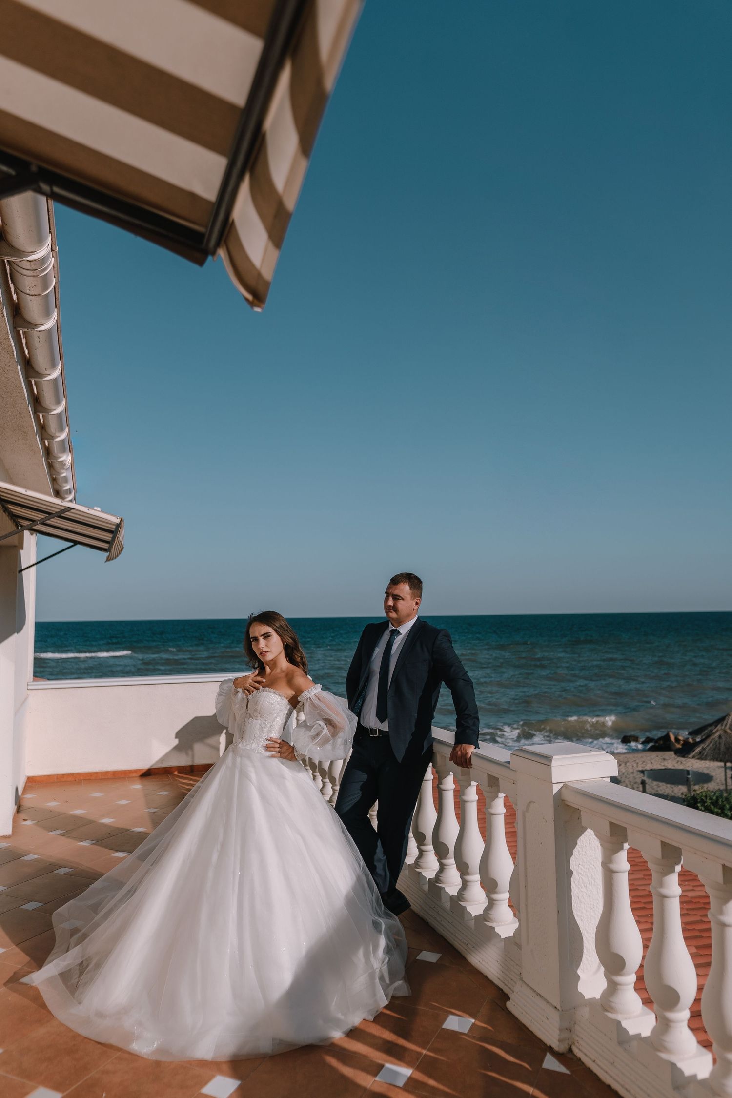 A wedding couple poses on an oceanfront balcony with white balustrades and stunning blue sea views on a sunny day.