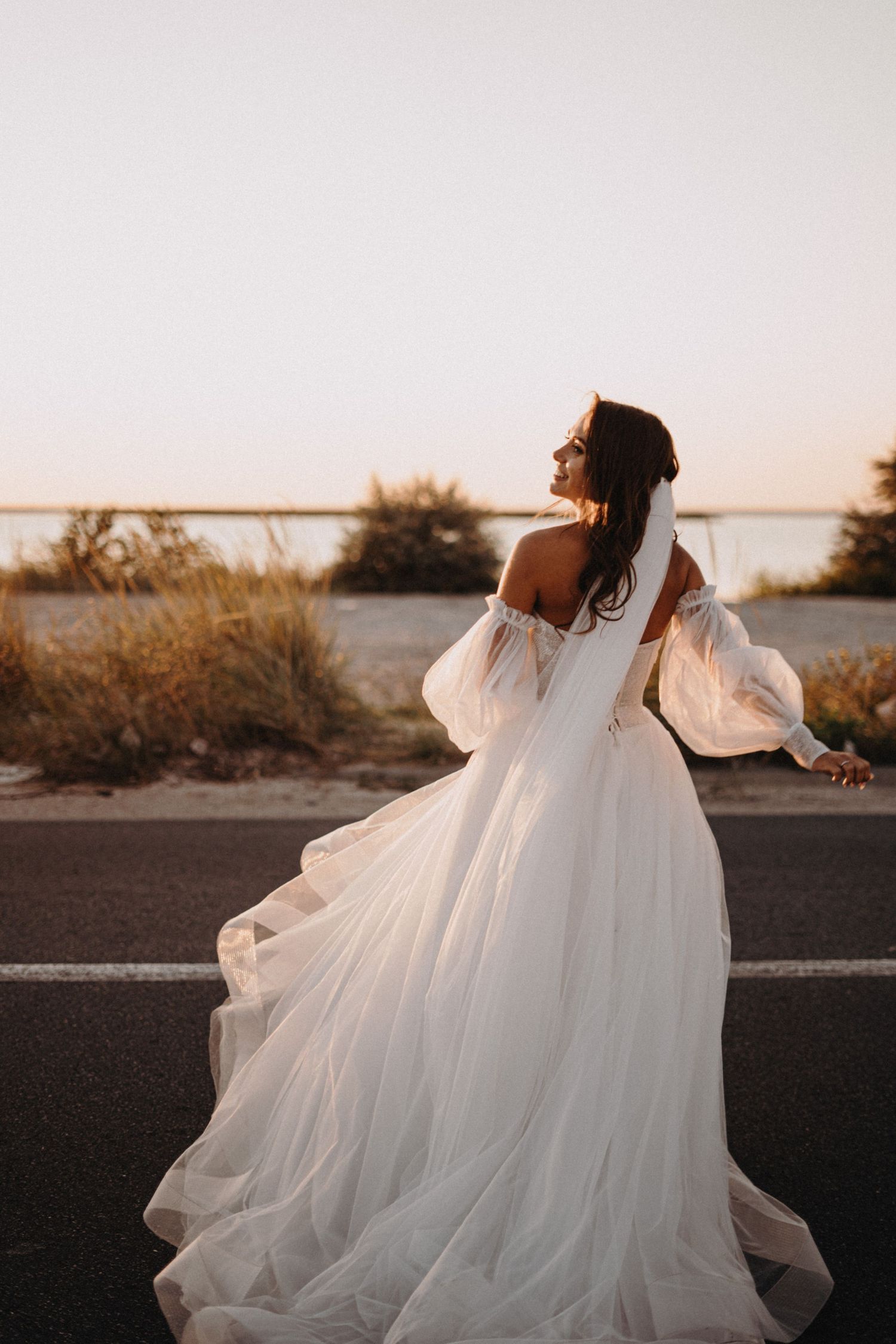 A bride in a flowing white gown twirls gracefully at sunset against a serene waterfront backdrop.