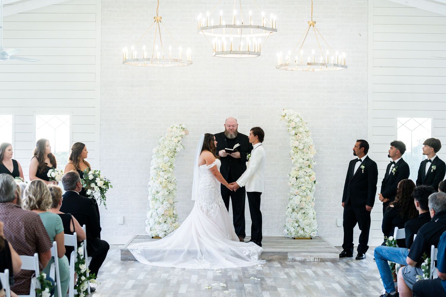 A wedding ceremony takes place under crystal chandeliers with white floral arches and guests looking on.