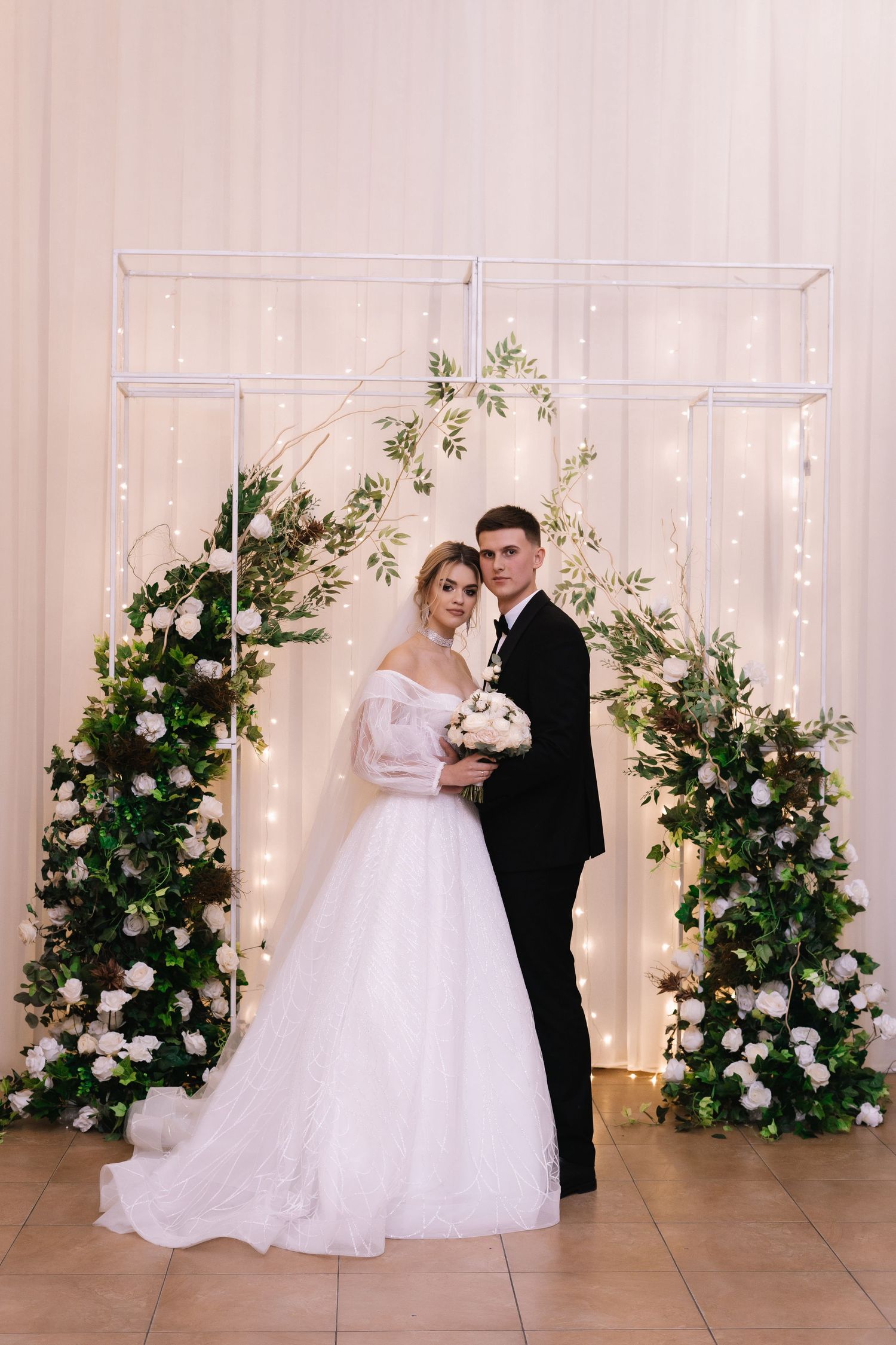 Wedding ceremony setup with floral arch made of white roses and greenery, with twinkling lights against white backdrop.
