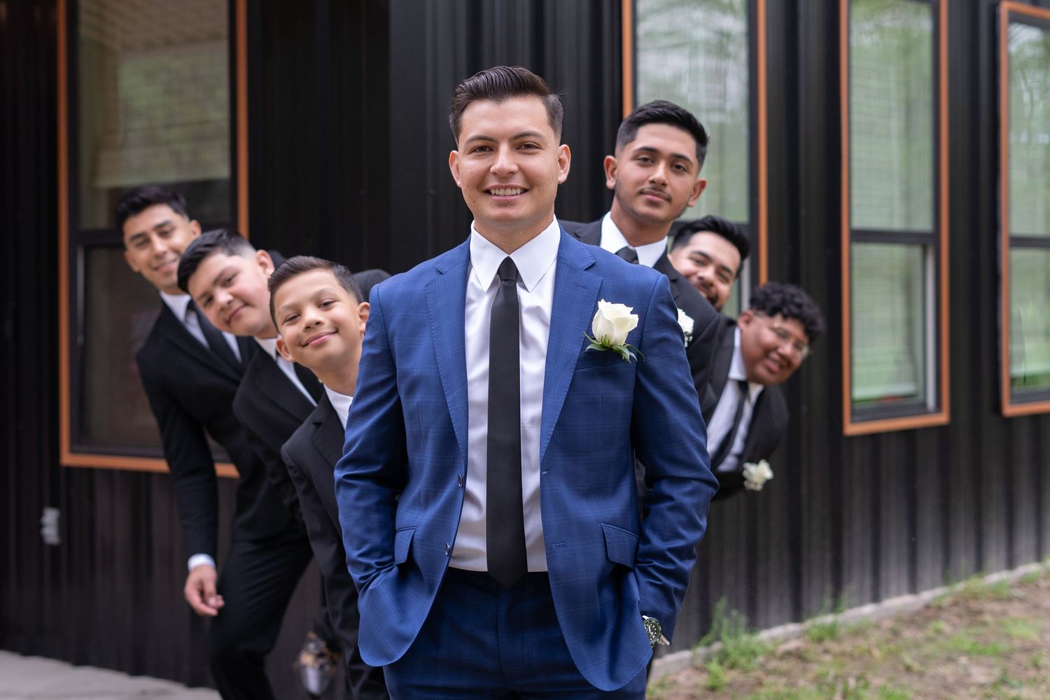 Groomsmen in navy blue suits pose playfully for wedding photos outside a rustic building.