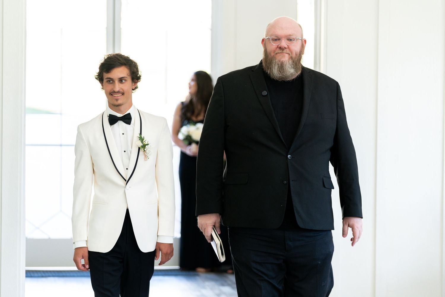 A group of people in formal black and white attire walk together down a white hallway.