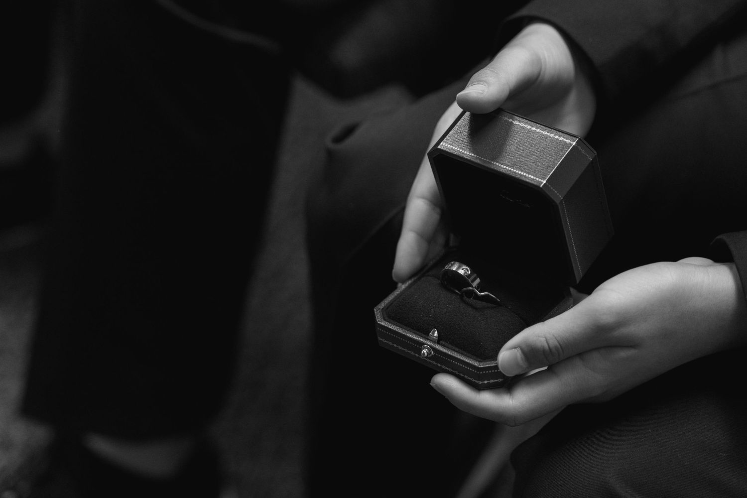 A black and white photo shows hands holding an open ring box with an engagement ring inside.