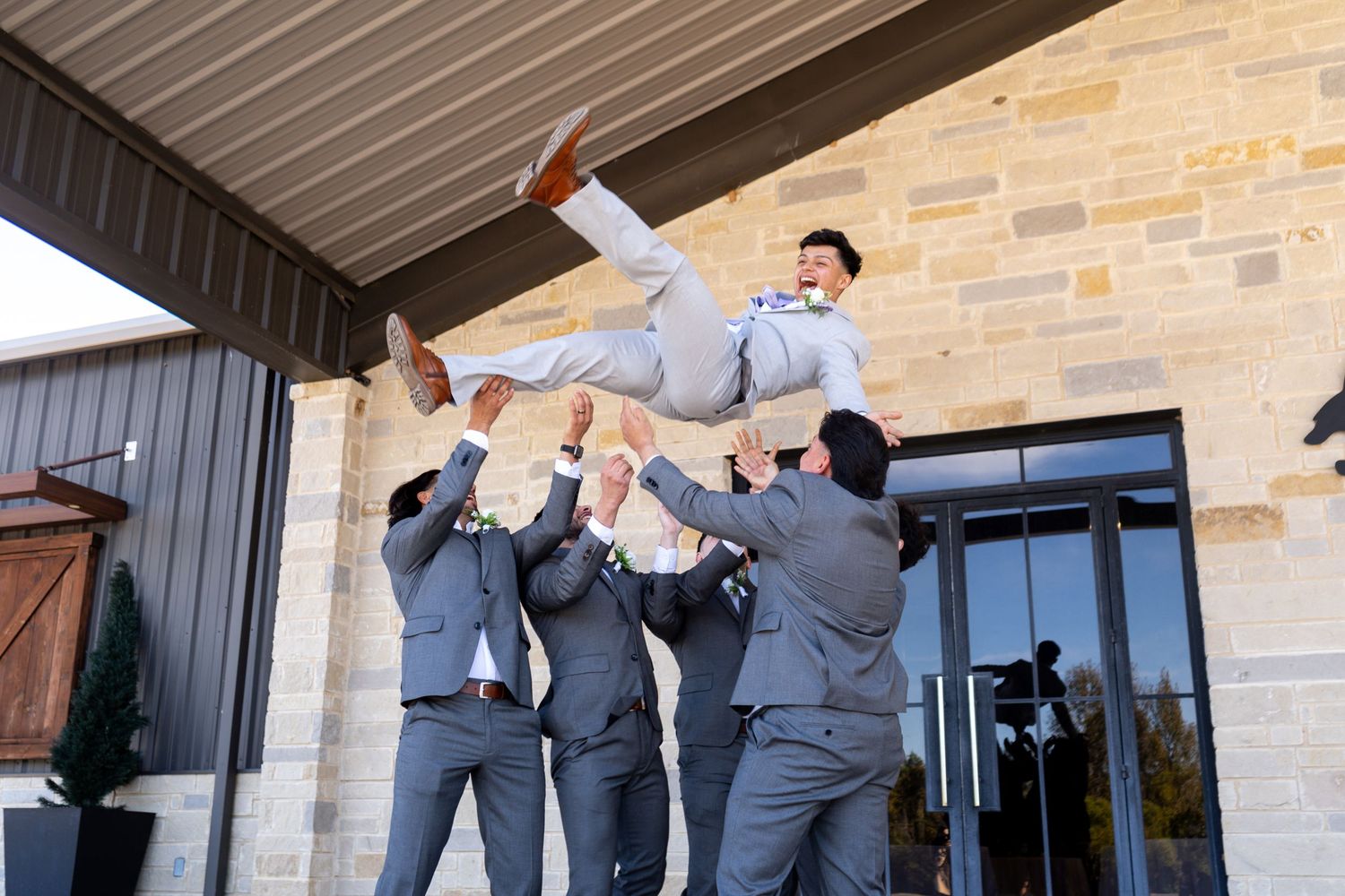 Groomsmen in gray suits jumping and posing playfully outside a stone building.