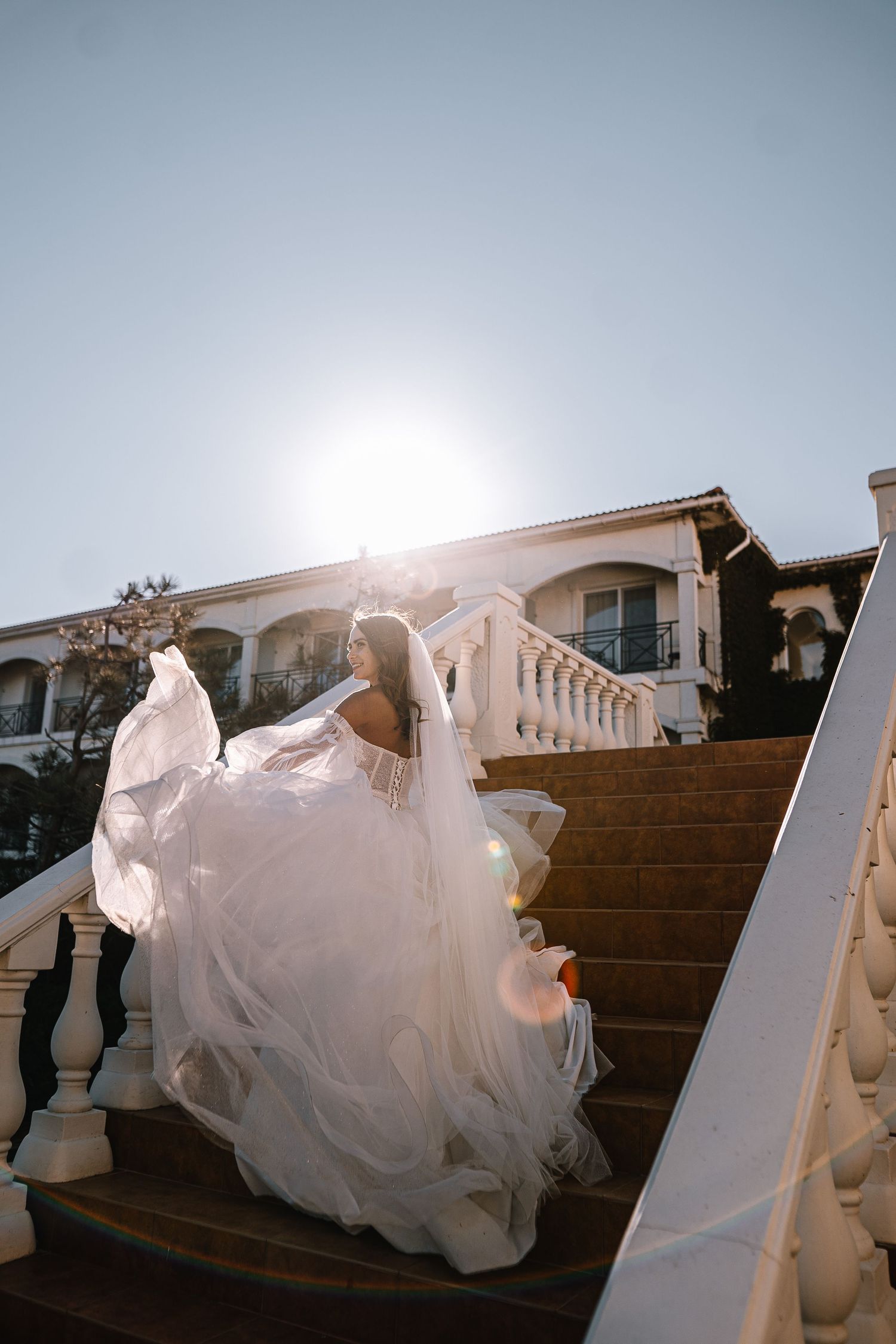 A flowing white wedding dress billows dramatically as the bride descends an elegant outdoor staircase at sunset.