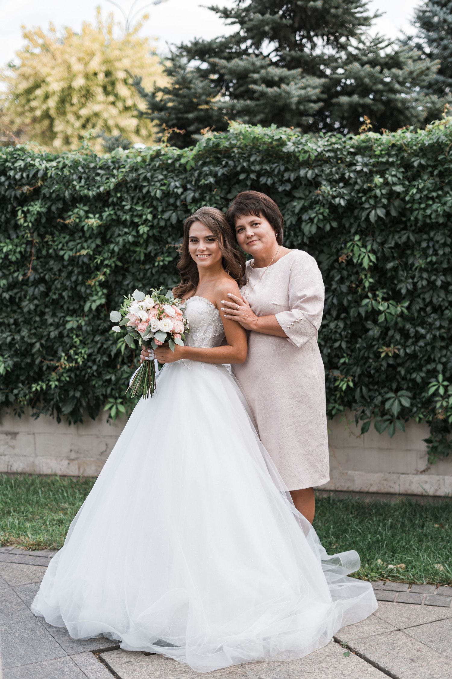 Two people sharing a joyful moment outdoors by evergreen shrubs during a wedding celebration.