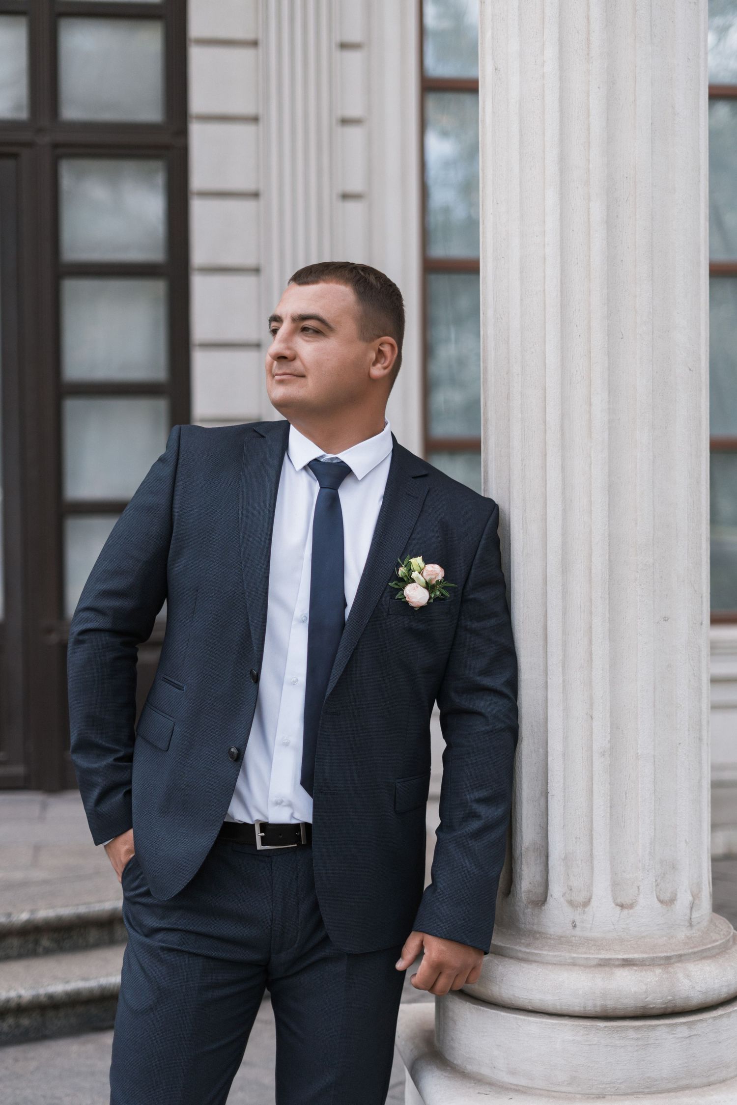 A groom in a dark suit with white boutonniere poses thoughtfully against a classical column at a wedding venue.