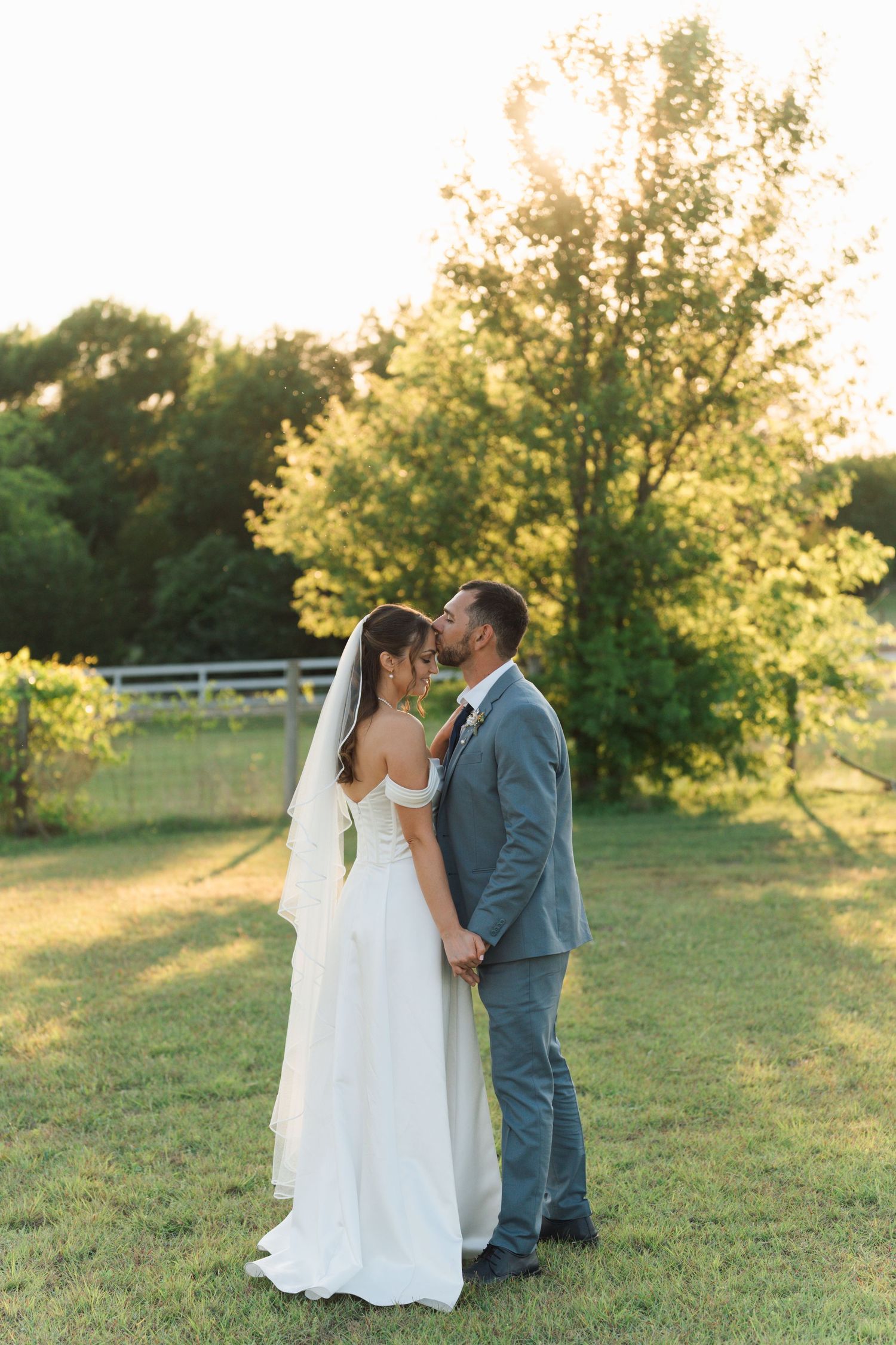 Bride and groom kissing outdoors at golden hour, surrounded by lush green trees on a sunny wedding day.