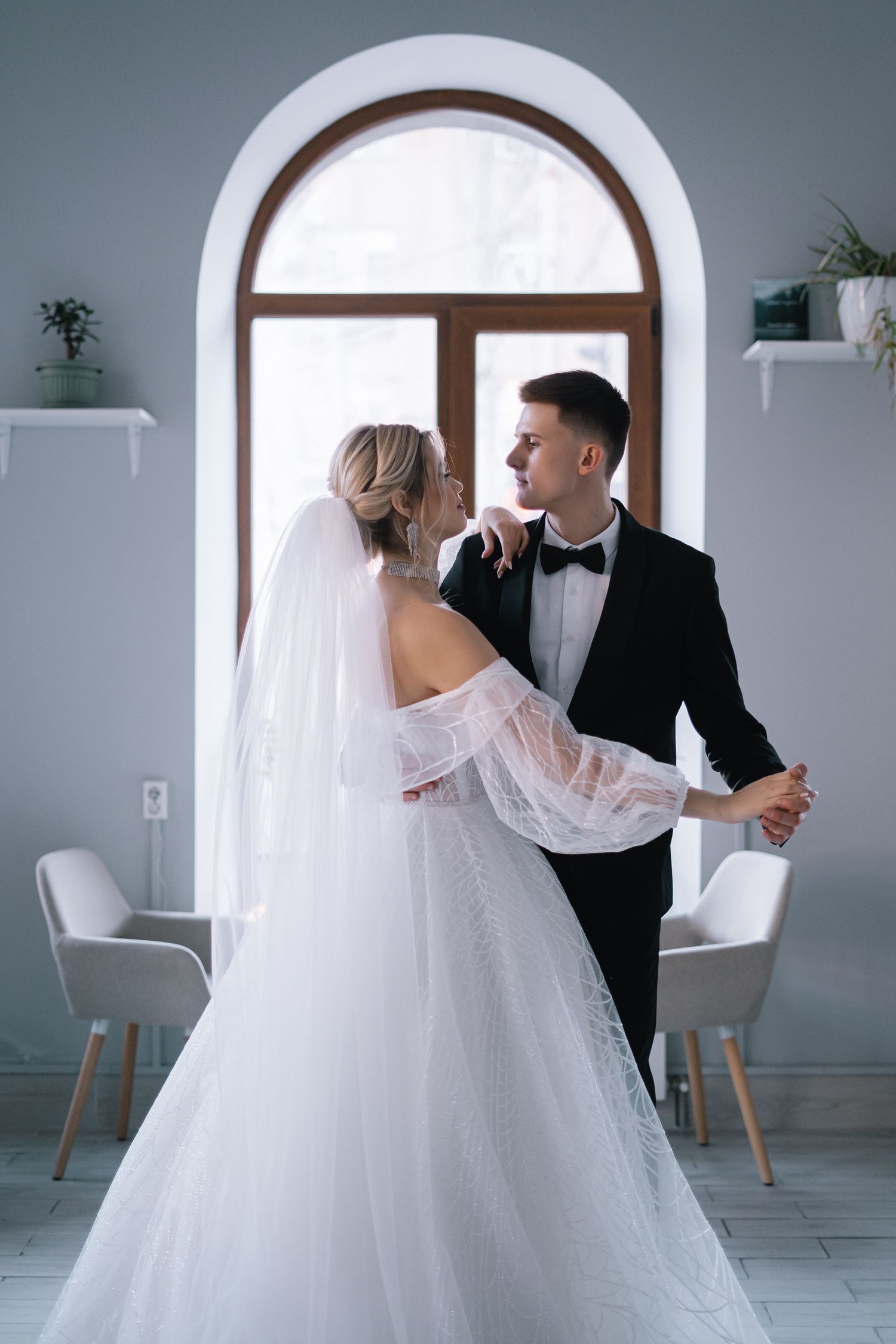 A romantic wedding scene with a couple in formal attire dancing together in an elegant room with arched windows.